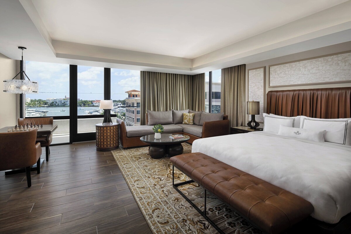 Une chambre d’hôtel moderne, dotée d’un grand lit, d’un canapé, d’une table basse et offrant une vue sur la ville grâce à de grandes fenêtres.