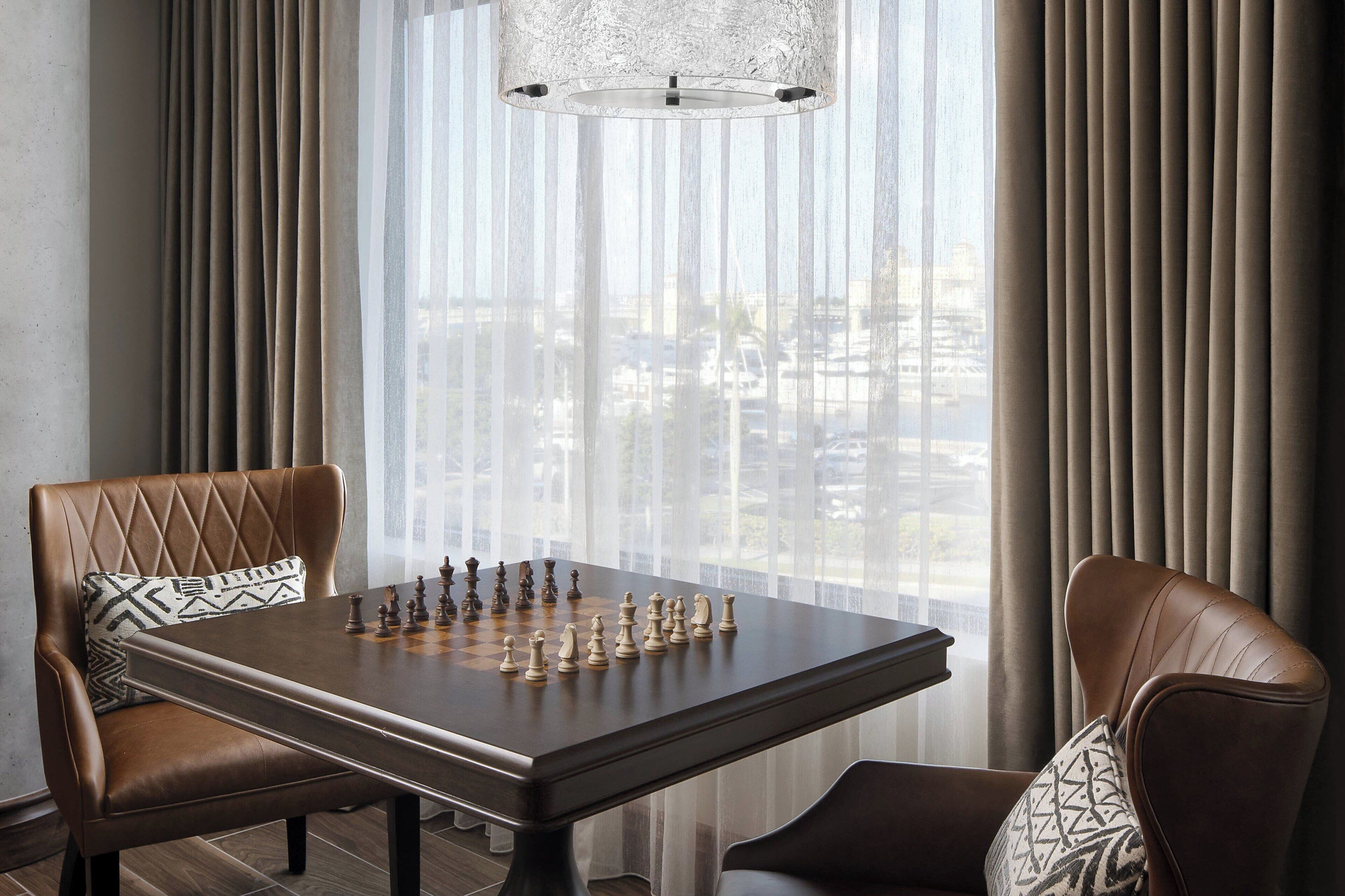 A chessboard set on a table in a room with two leather chairs, a patterned pillow, and a view through sheer curtains.