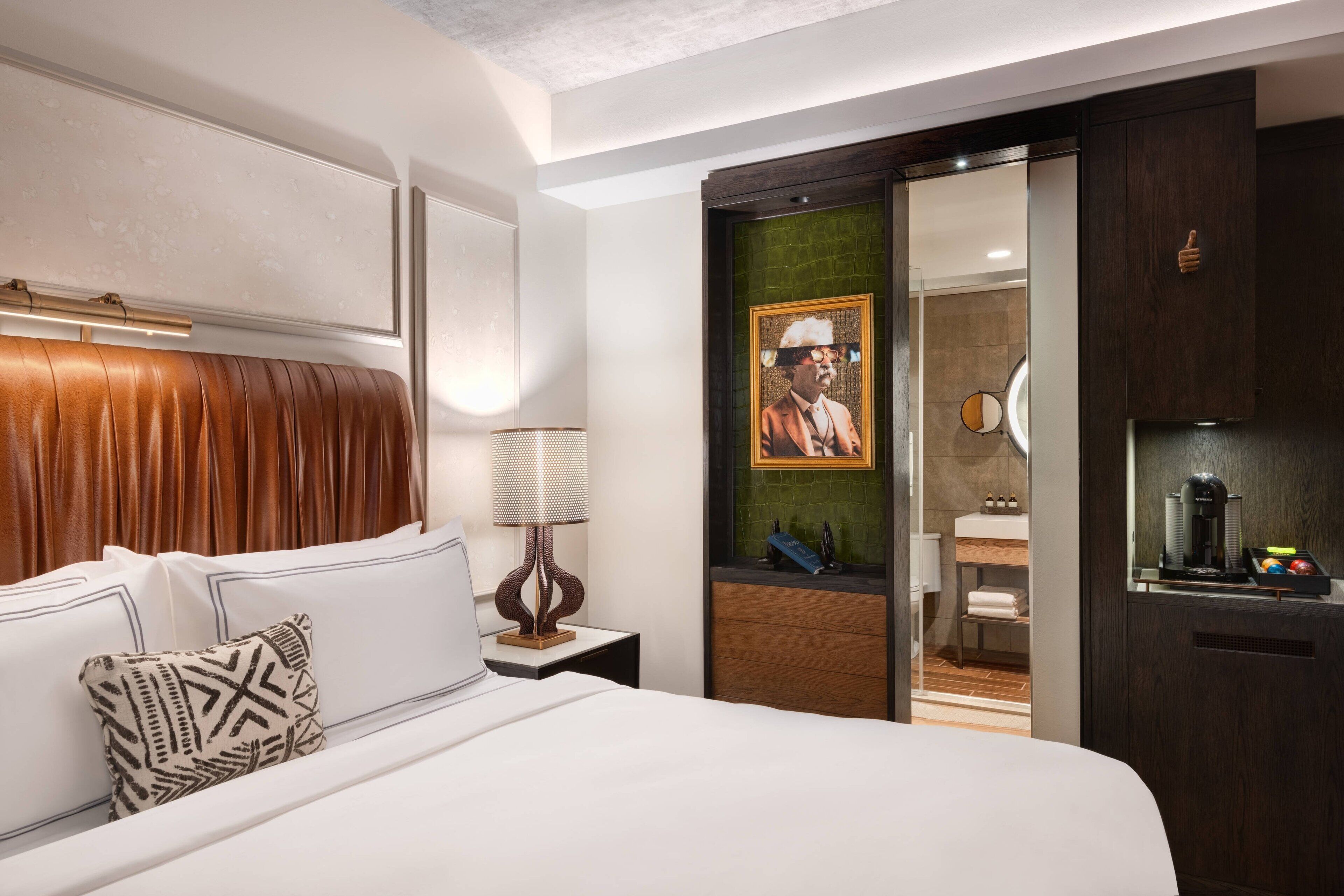 Une chambre d’hôtel moderne avec un grand lit, une tête de lit en bois, une table de chevet avec une lampe, un tableau au mur et une vue sur la salle de bain.