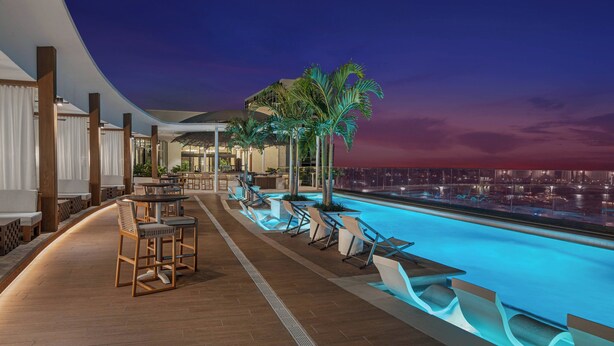 Un área de piscina en la azotea con sillones y mesas, palmeras, y vistas de la ciudad al atardecer.