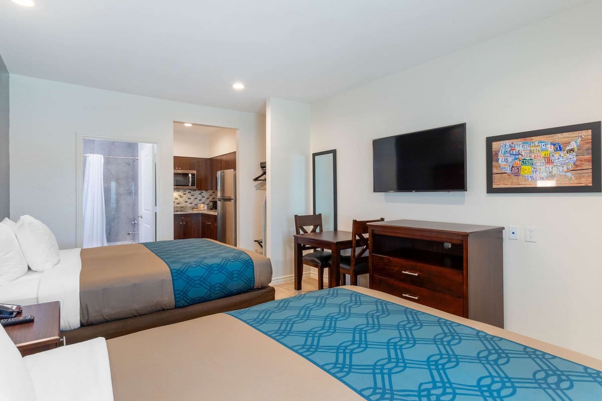 A hotel room with two beds, a desk, a chair, a TV, and a kitchen area in the background.