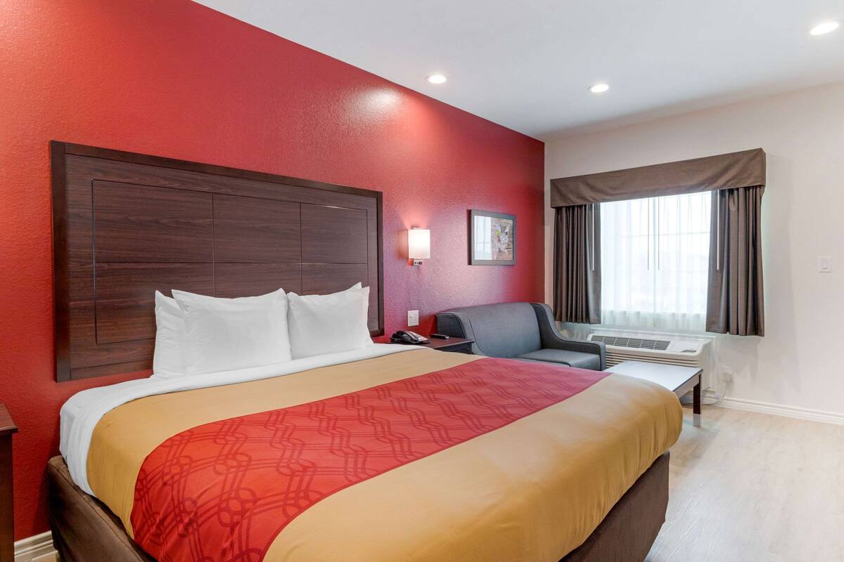 A hotel room with a large bed, a desk, a chair, a wall-mounted air conditioning unit, and a window with curtains.