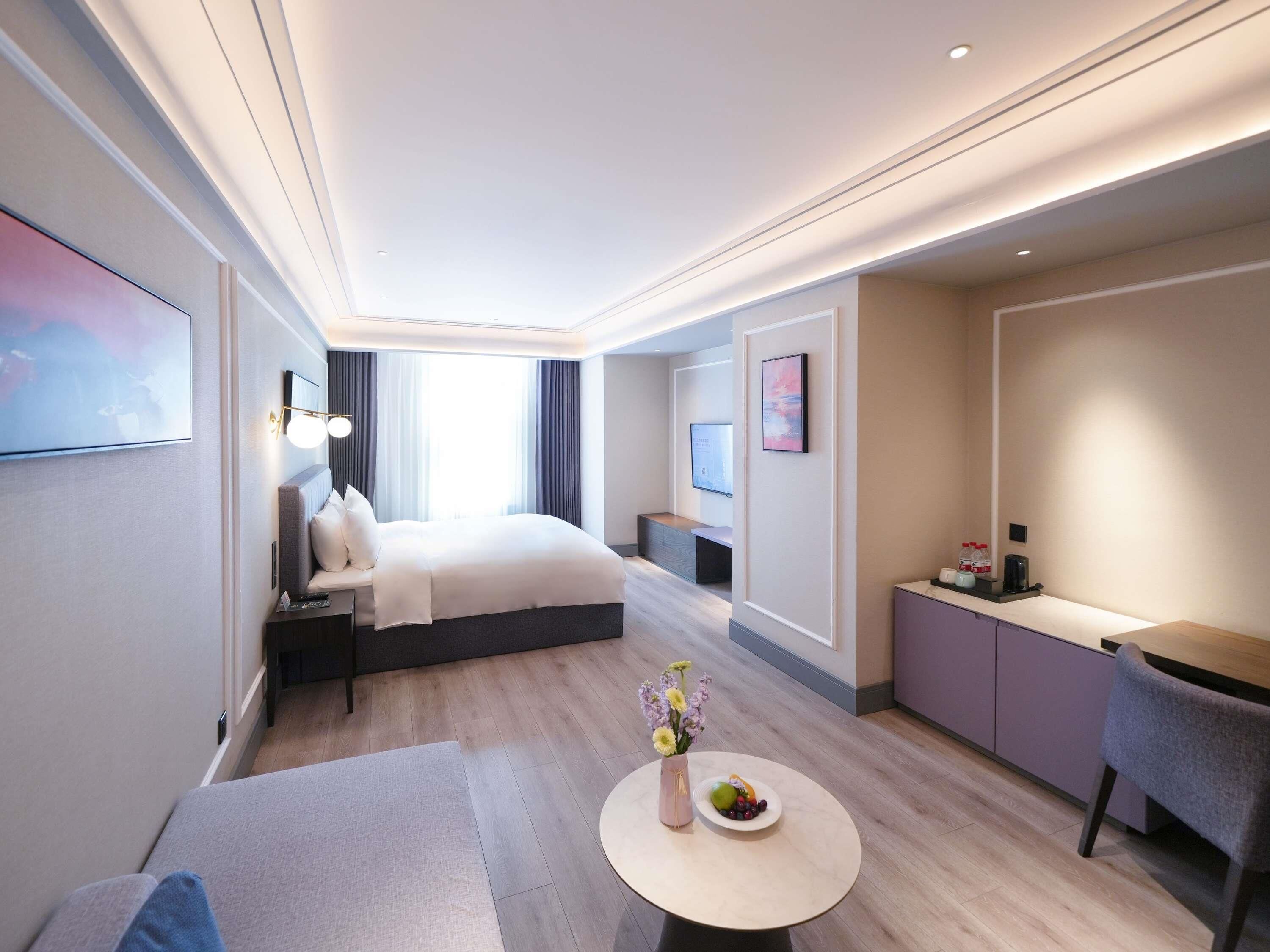 A modern hotel room with a bed, bedside table, lamp, and a small round table with a vase and fruit.