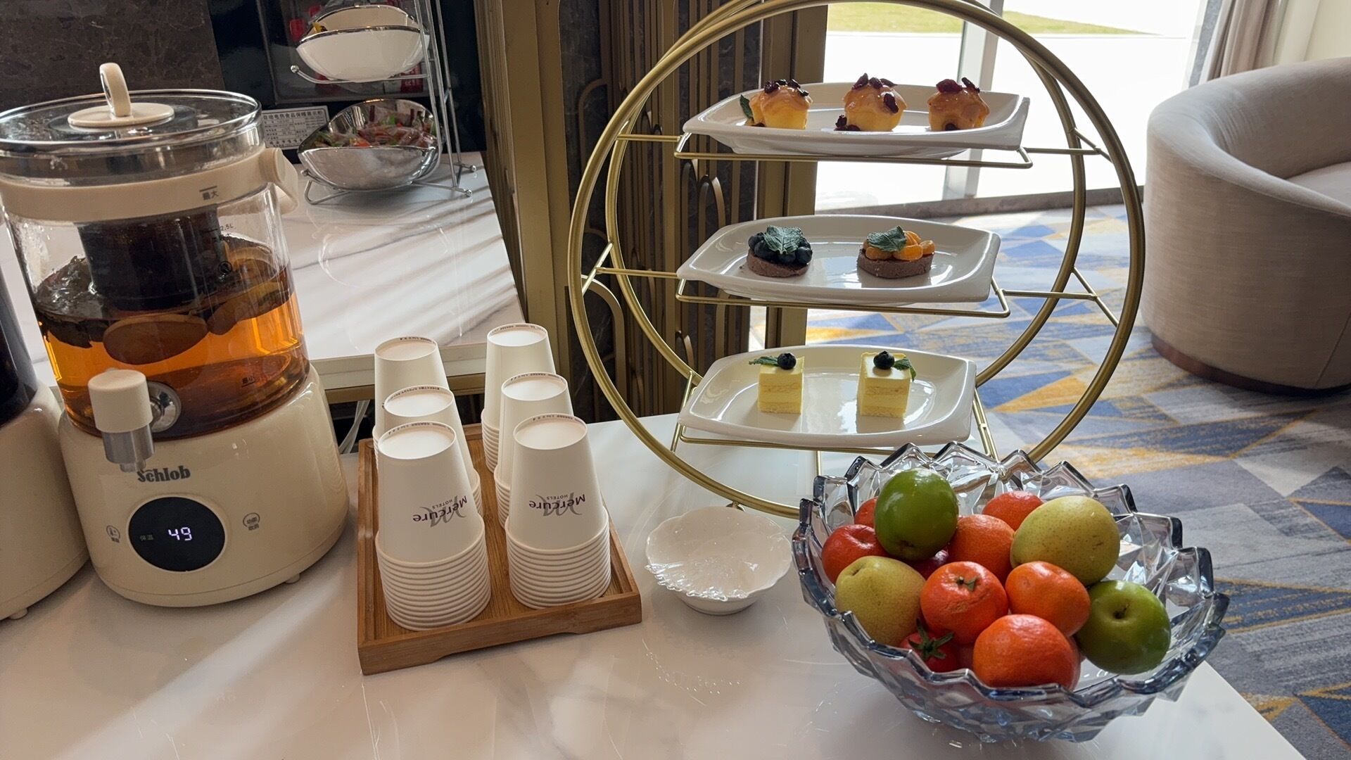 A buffet setup with a coffee machine, stacked plates, a fruit bowl, and tiered dessert plates.
