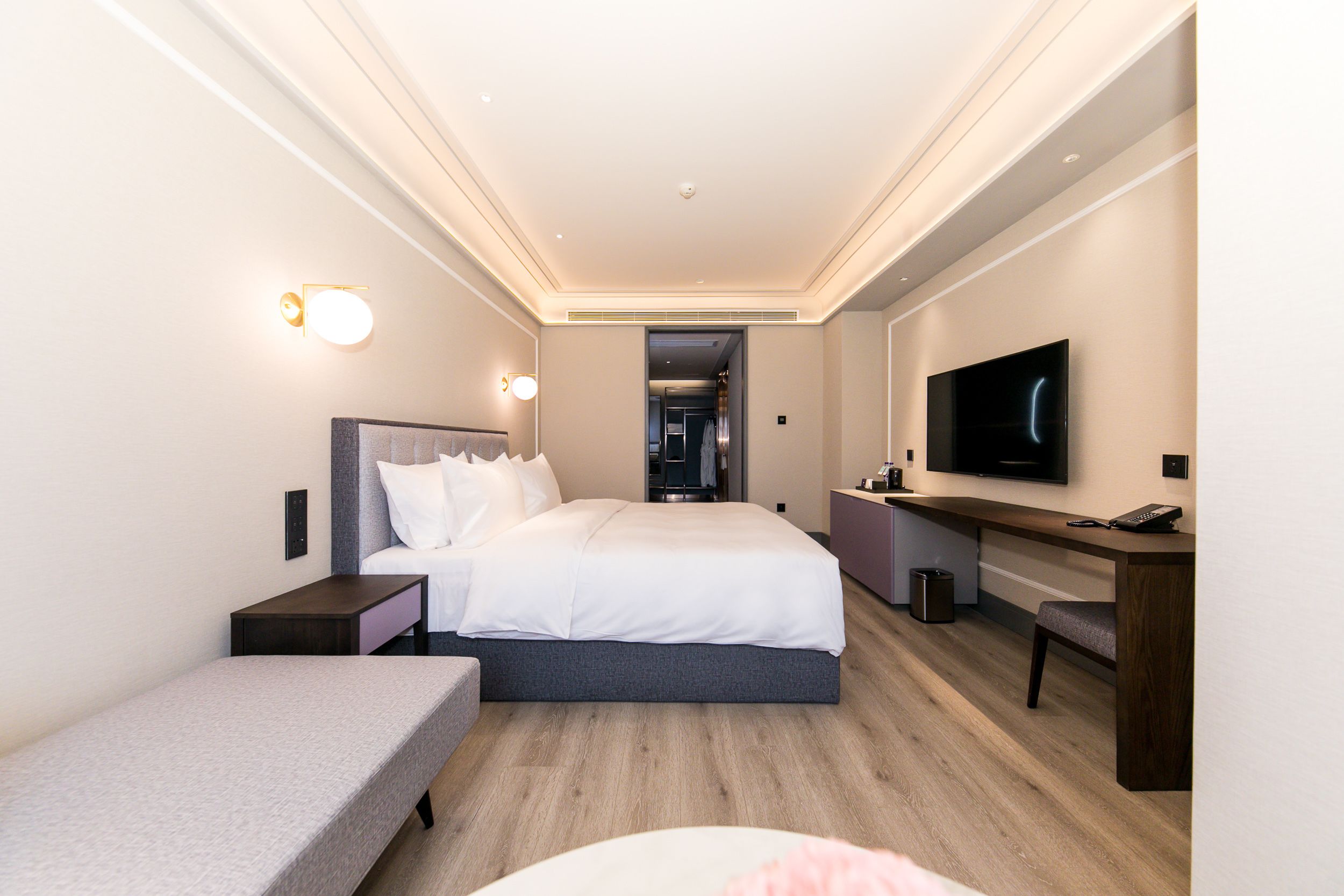 A modern hotel room with a large bed, a desk, a TV, and a bench.