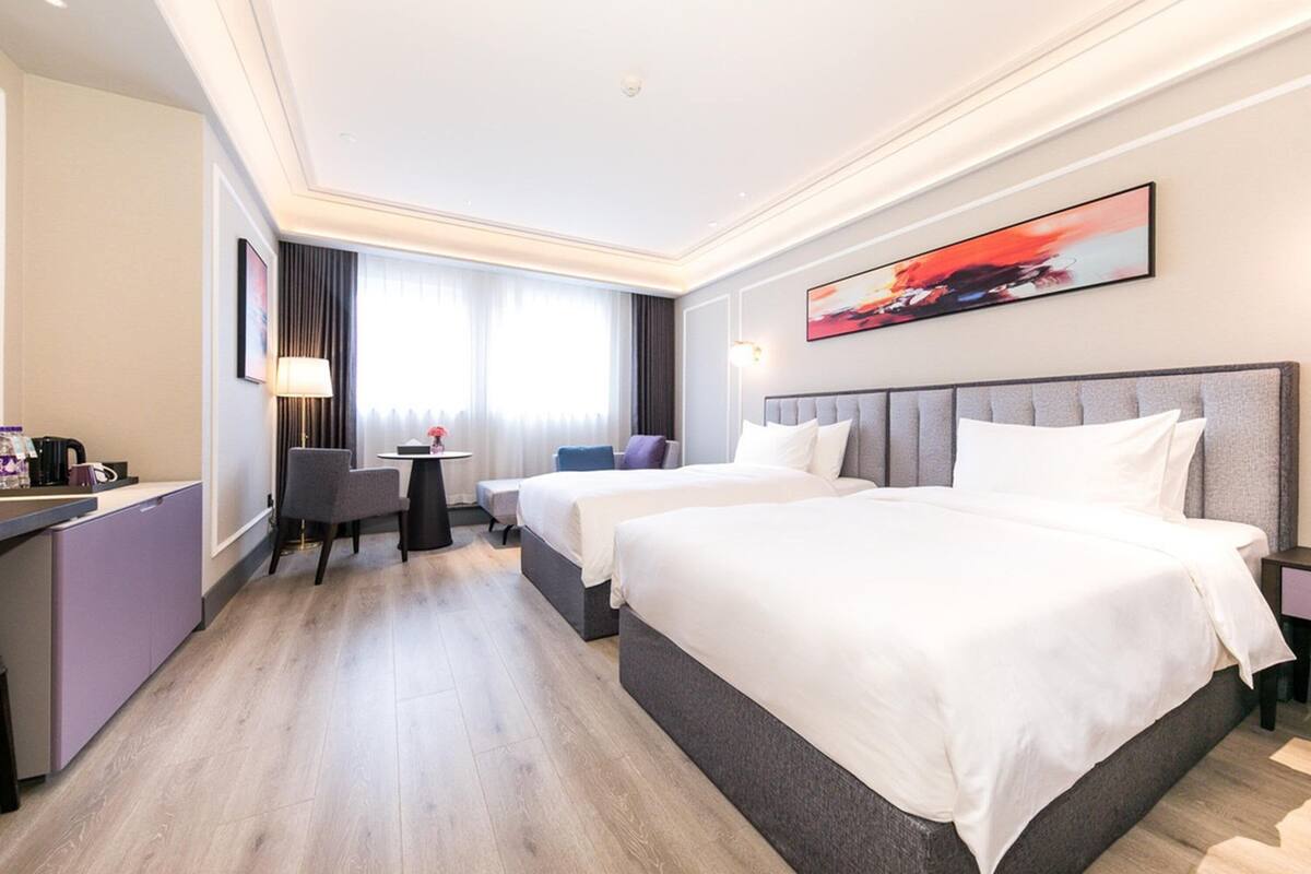 A hotel room with two beds, a desk, a chair, a small table, and a painting on the wall.