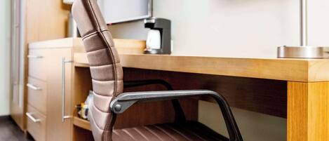 Un fauteuil en cuir marron, doté d’un haut dossier et d’accoudoirs, est placé devant un bureau en bois, sur lequel se trouvent une machine à café et un tableau encadré accroché au mur.