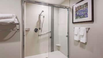 A glass-enclosed shower with a handheld showerhead and a handheld faucet. There are white towels on a rack and a framed picture on the wall.