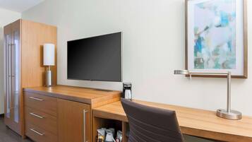 A hotel room with a wooden desk, a flat-screen TV, a coffee maker, a lamp, a framed abstract painting, and a brown chair.