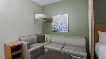 A modern hotel room with a grey sectional sofa, a wooden side table, a brown ottoman, a framed abstract painting, and a bed with white linens.