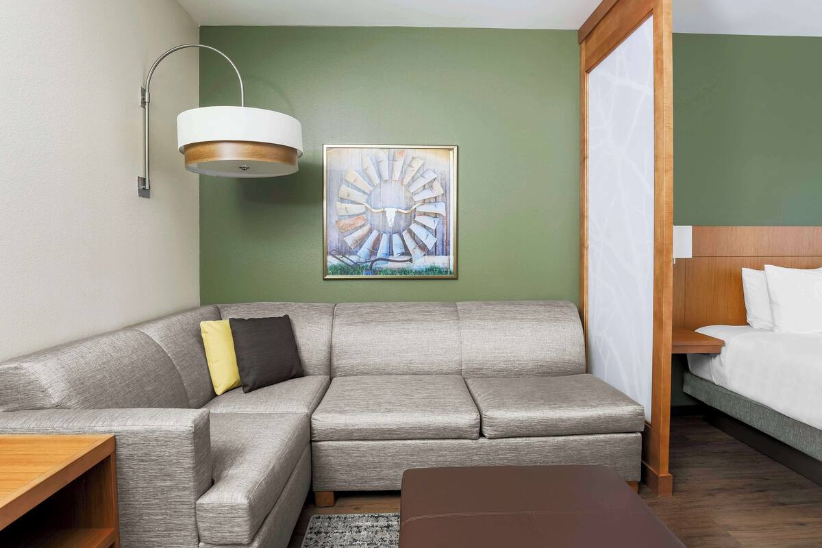 A modern hotel room with a grey sectional sofa, a wooden side table, a brown ottoman, a large abstract painting, and a bed with white linens.