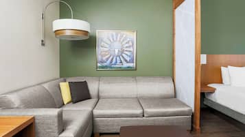 A modern hotel room with a grey sectional sofa, a wooden side table, a brown ottoman, a large abstract painting, and a bed with white linens.