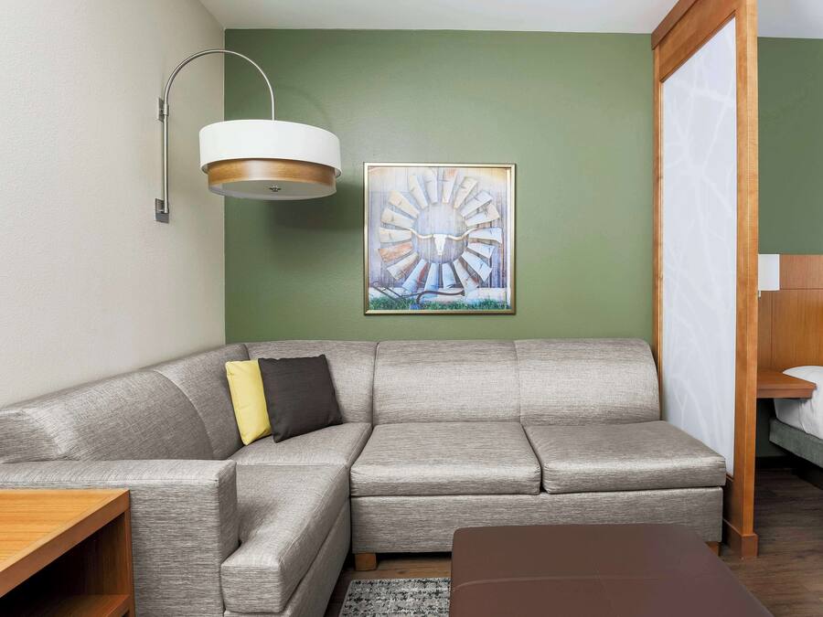 Una habitación de hotel moderna con un sofá seccional gris, una mesita de madera, un reposapiés marrón, un cuadro abstracto grande y una cama con ropa de cama blanca.