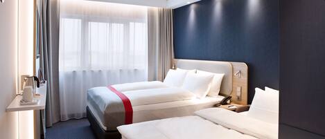A hotel room with two beds, a desk with a lamp, and a window with curtains.