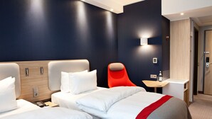 A hotel room with two beds, a red chair, a small table with a bottle, and a wall-mounted light.