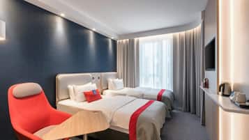 A hotel room with a curved ceiling, two beds with red accents, a red armchair, a bedside table with a lamp, and a TV on the wall.
