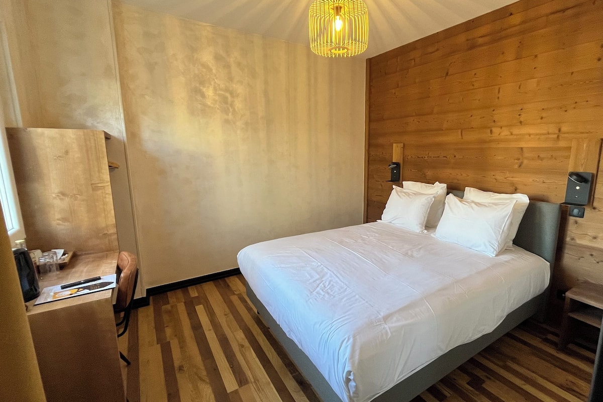 A hotel room with a wooden headboard, a bed with white linens, a wooden floor, and a hanging light fixture.