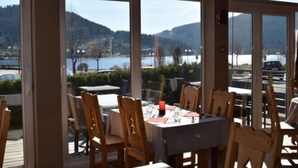 Un espacio de comedor al aire libre con mesas y sillas, grandes ventanales que ofrecen vistas a un lago y montañas.