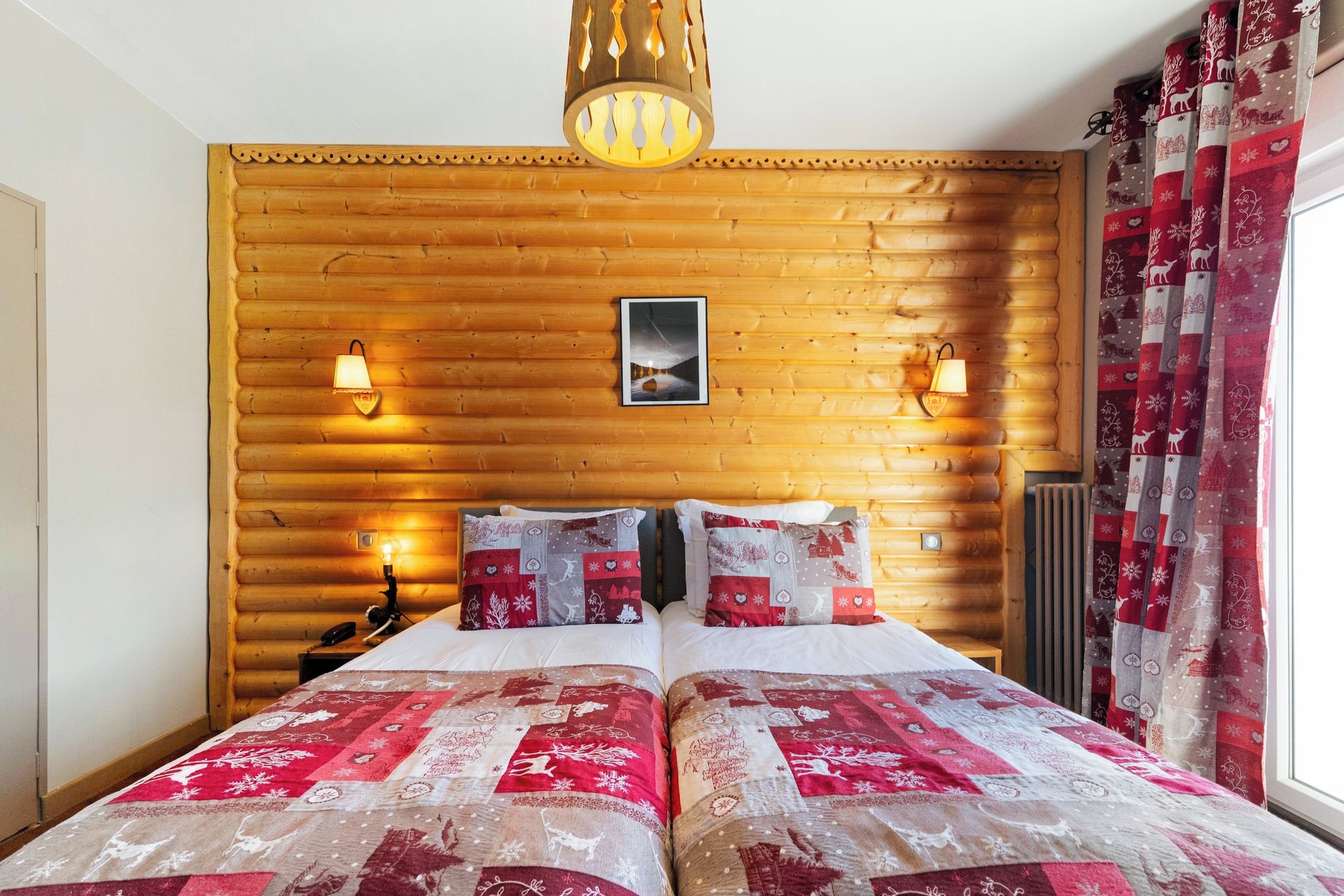 A room with a wooden log wall, a bed with a red and white quilt, two wall-mounted lamps, and a picture on the wall.