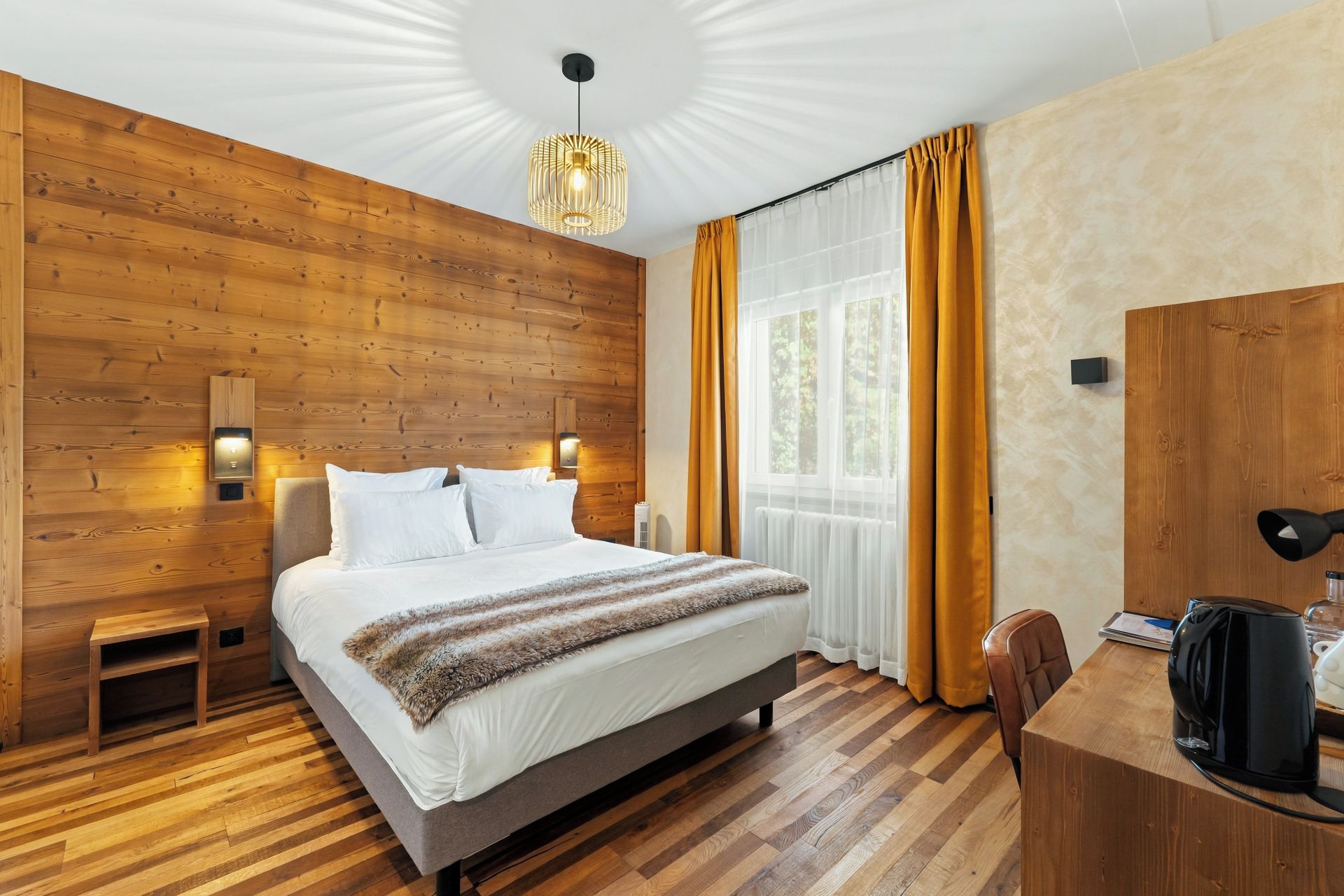 Un dormitorio moderno con cabecera de madera, cama con ropa de cama blanca, una mesita de noche pequeña, un escritorio con cafetera y un ventanal con cortinas.