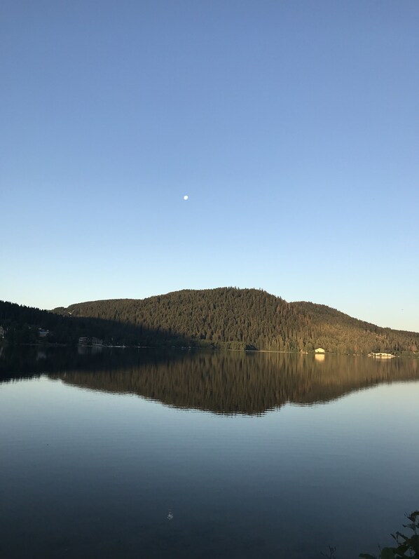 A calm lake with a clear reflection of a forested hill and a small building on the shore. - L’Annexe by La Réserve (Gerardmer)