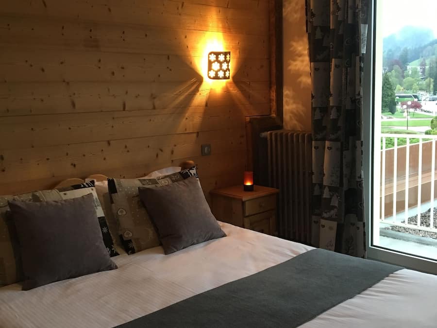 A wooden room with a bed, a nightstand, a wall-mounted lamp, and a view of the outdoors.