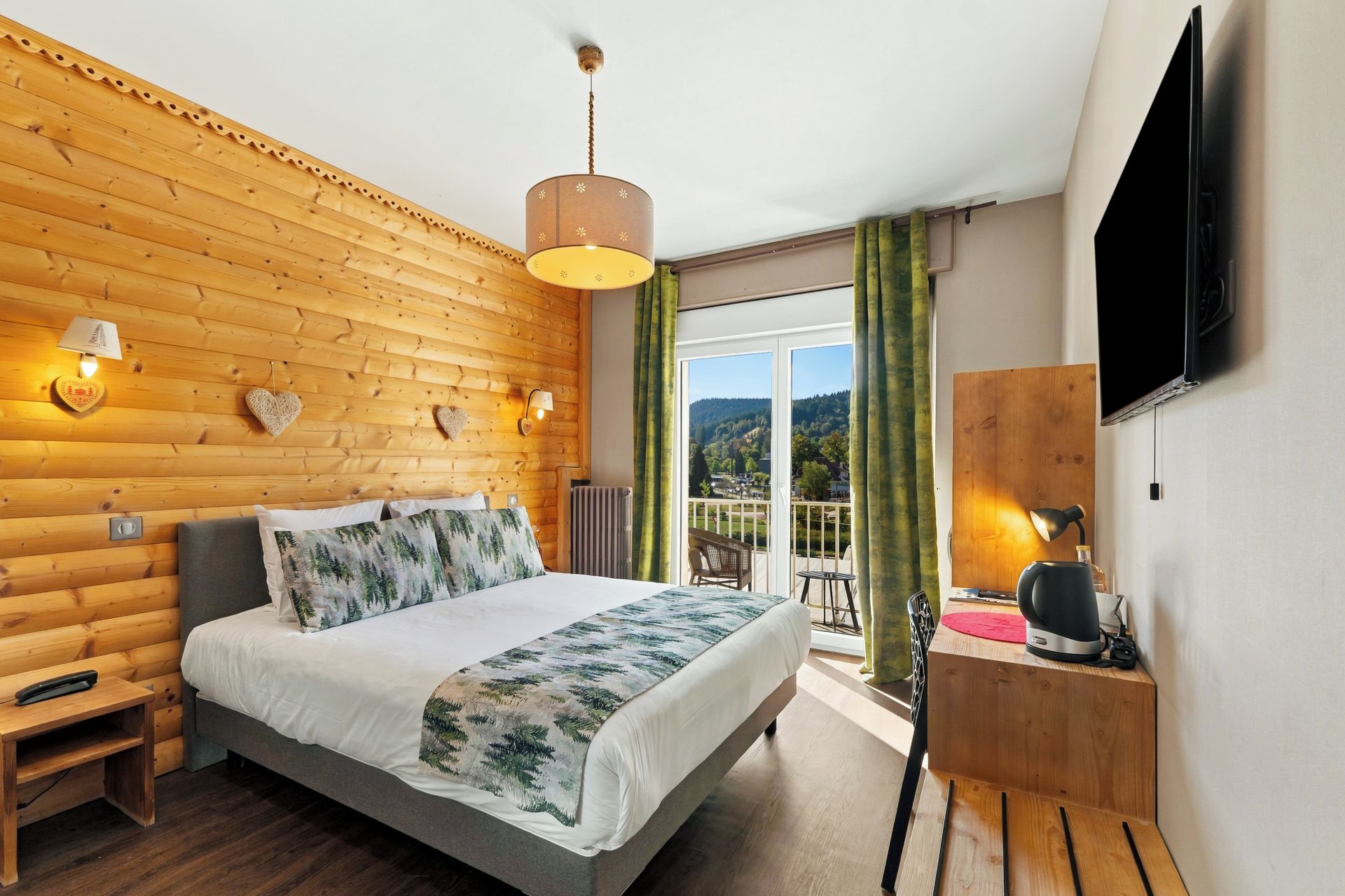 Une chambre d’hôtel avec un mur en bois, un lit recouvert d’une courtepointe à motifs forestiers, un balcon avec vue et un téléviseur à écran plat fixé au mur.