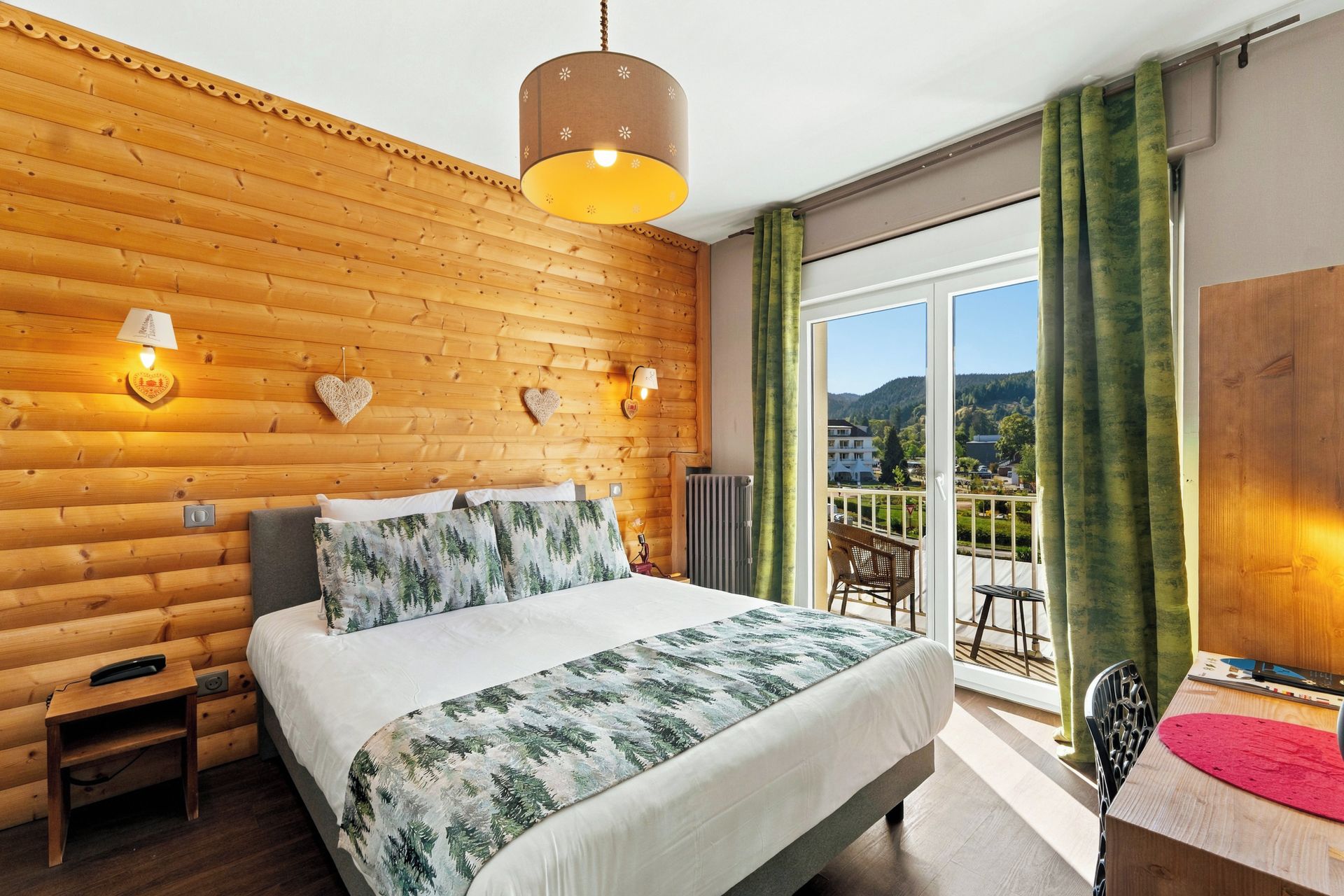 Habitación de hotel con pared de madera rústica, cama con cubrecama de motivos forestales, balcón con vistas al campo y lámpara colgante.