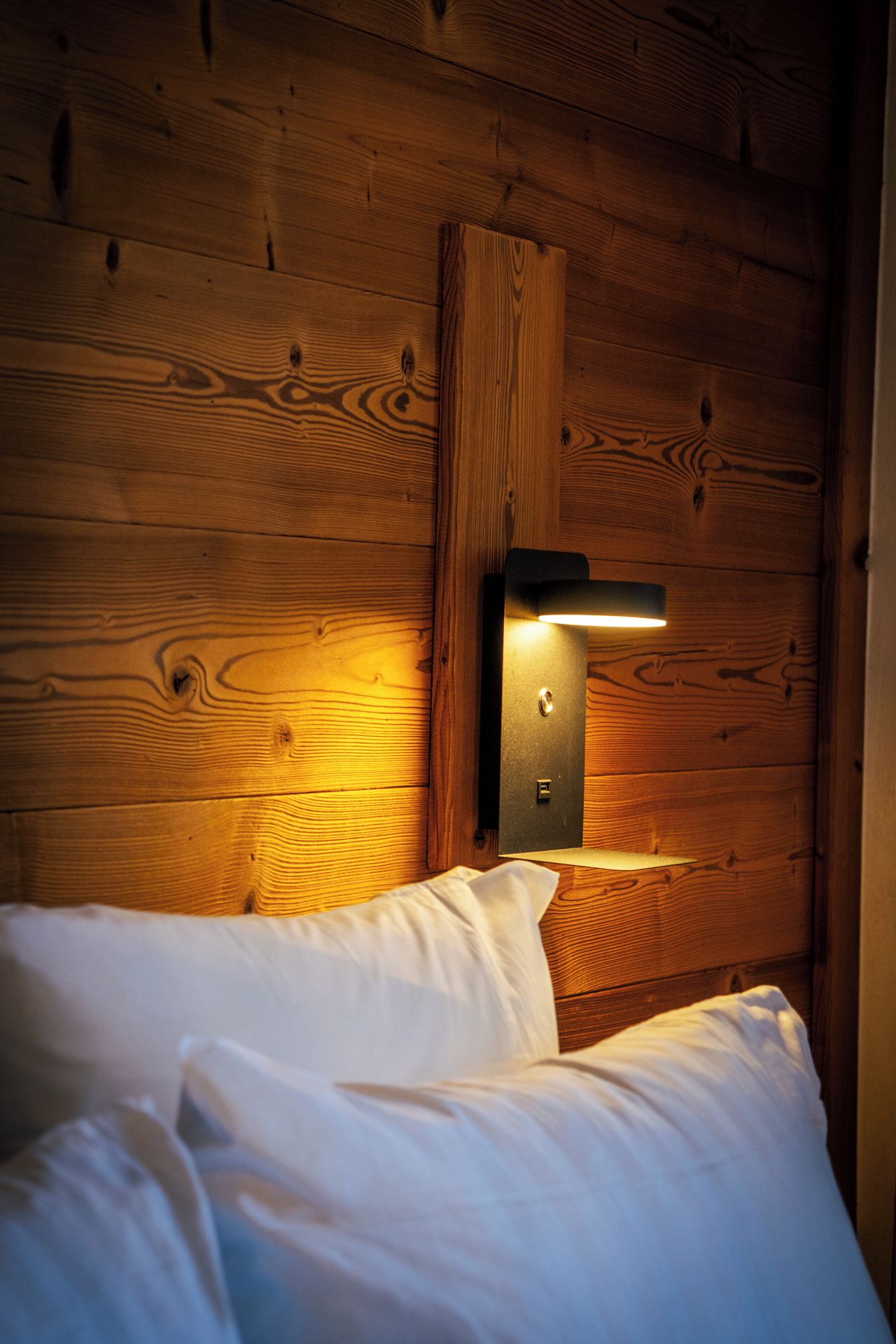 Pared de madera con un aplique integrado sobre una cama con ropa blanca.
