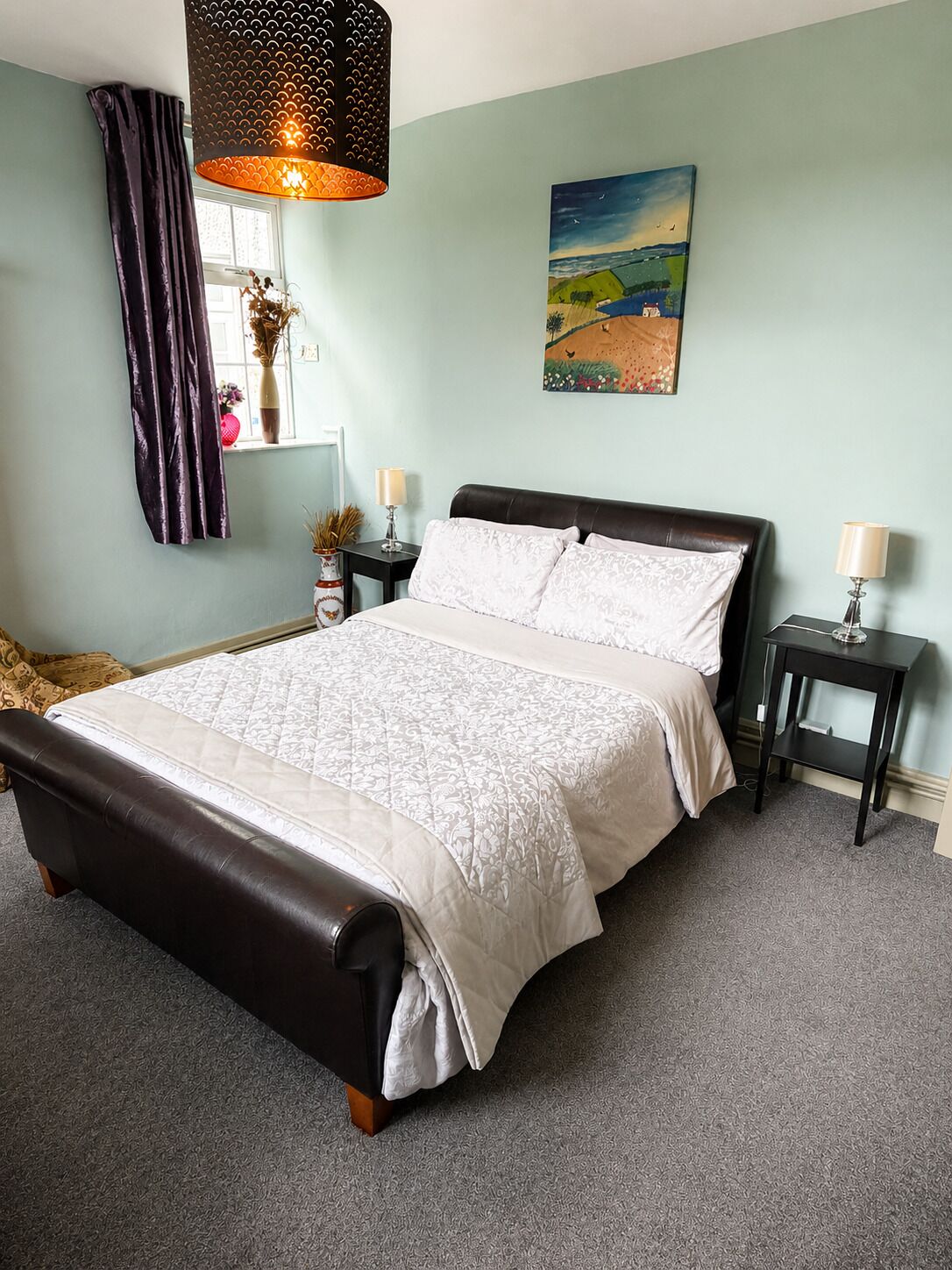 A bedroom with a bed, two bedside tables, a window with curtains, a painting on the wall, and a hanging light fixture.