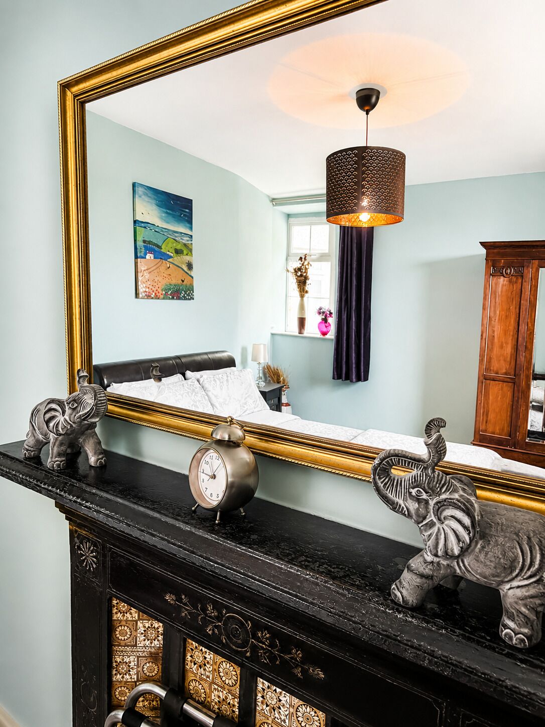 A room with a large mirror, a fireplace with decorative elephants, a clock, and a painting on the wall.