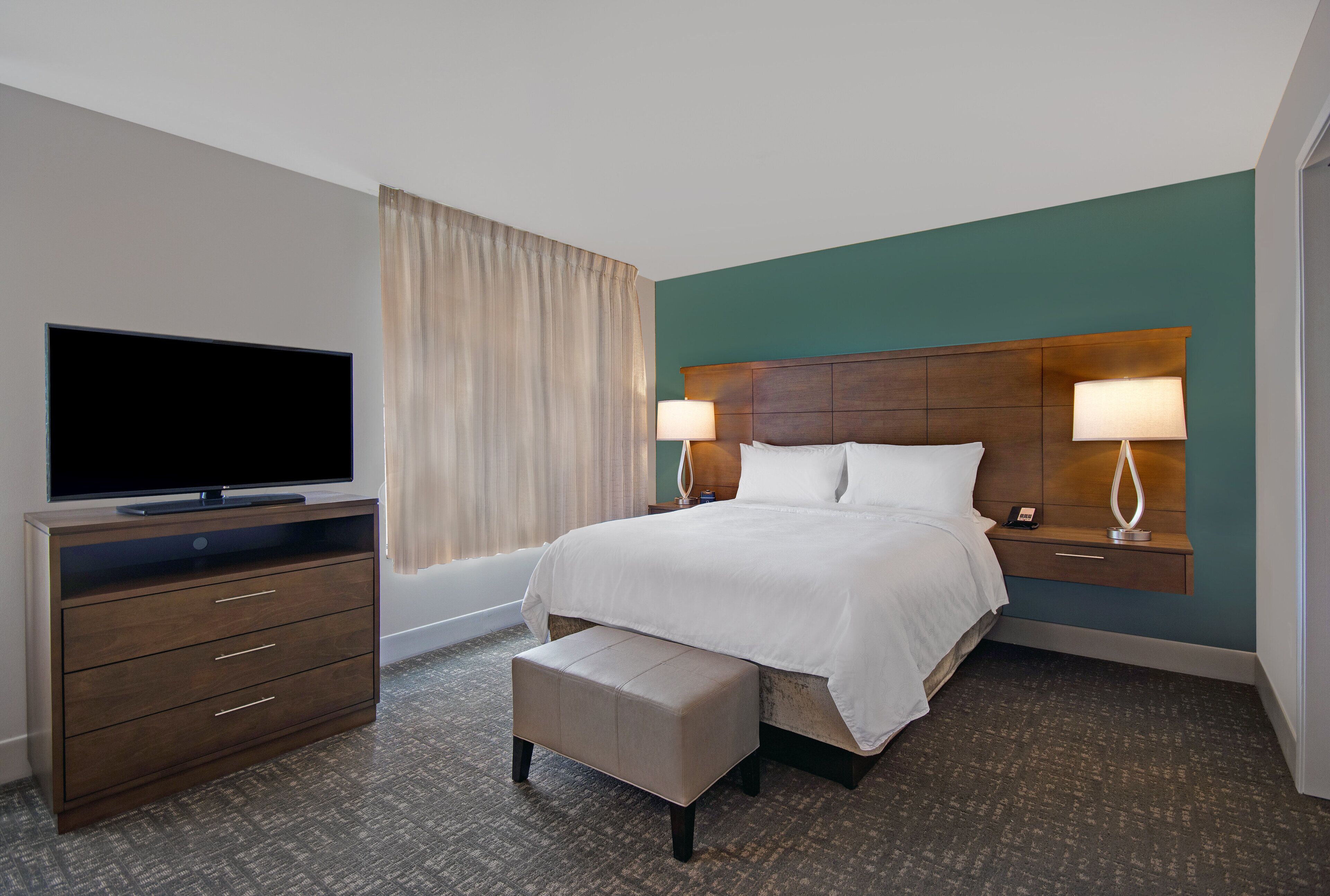 Quarto de hotel com uma cama, televisão em uma cômoda, mesa de cabeceira com abajur e janela com cortinas.