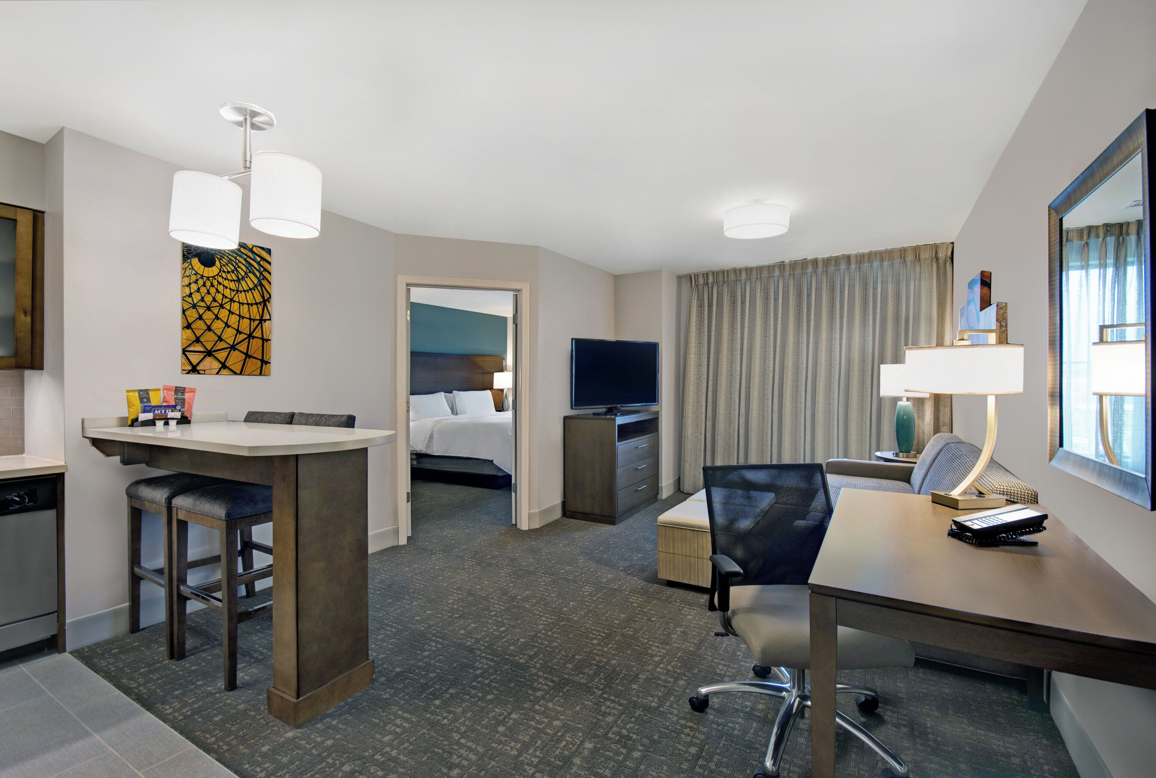 A hotel room with a kitchenette, a bed, a TV, a desk with a chair, and a window with curtains.