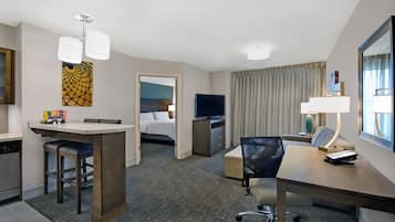 A hotel room with a kitchenette, a bed, a TV, a desk with a chair, and a window with curtains.