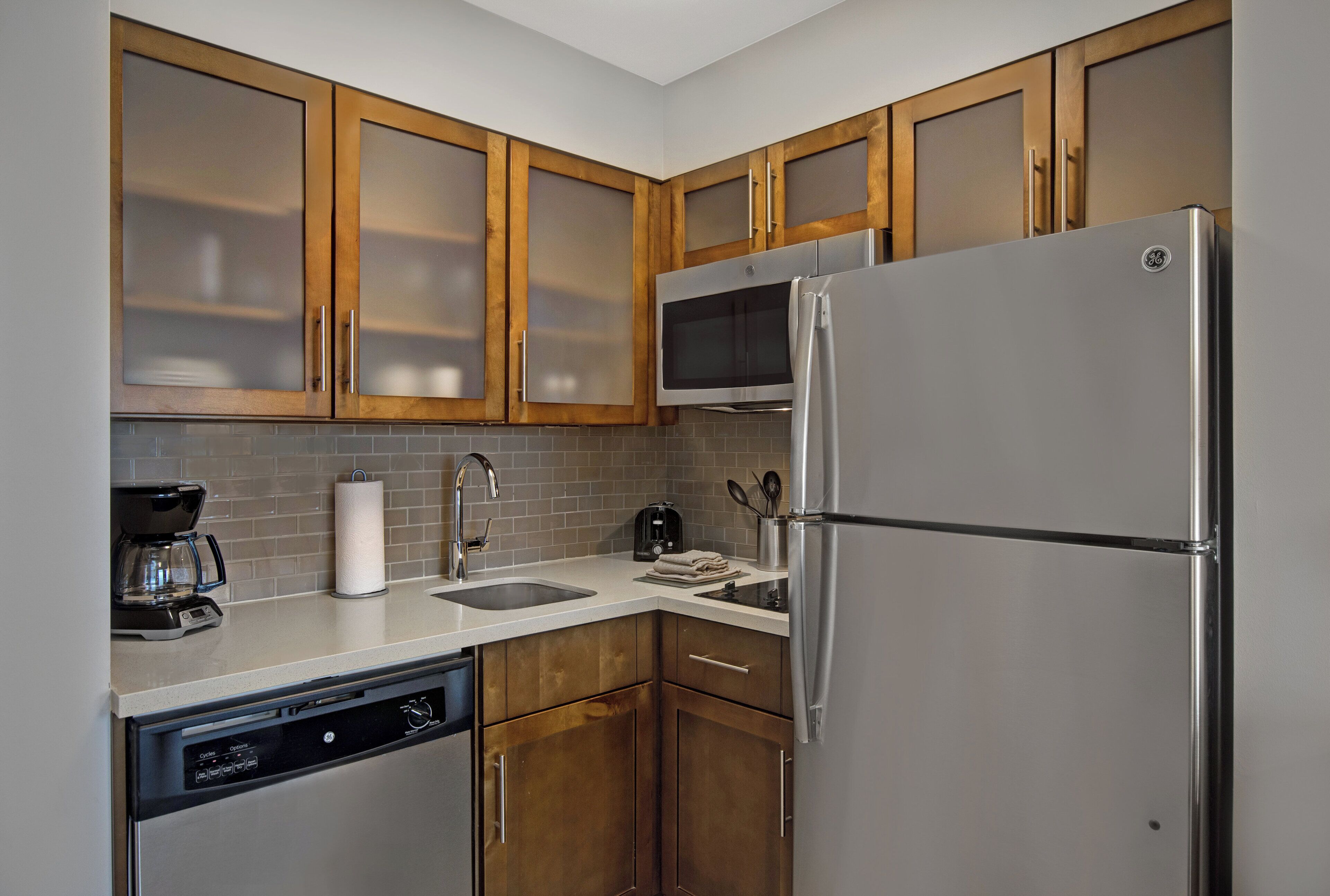 A compact kitchen with wooden cabinets, a stainless steel refrigerator, a microwave, and a dishwasher.