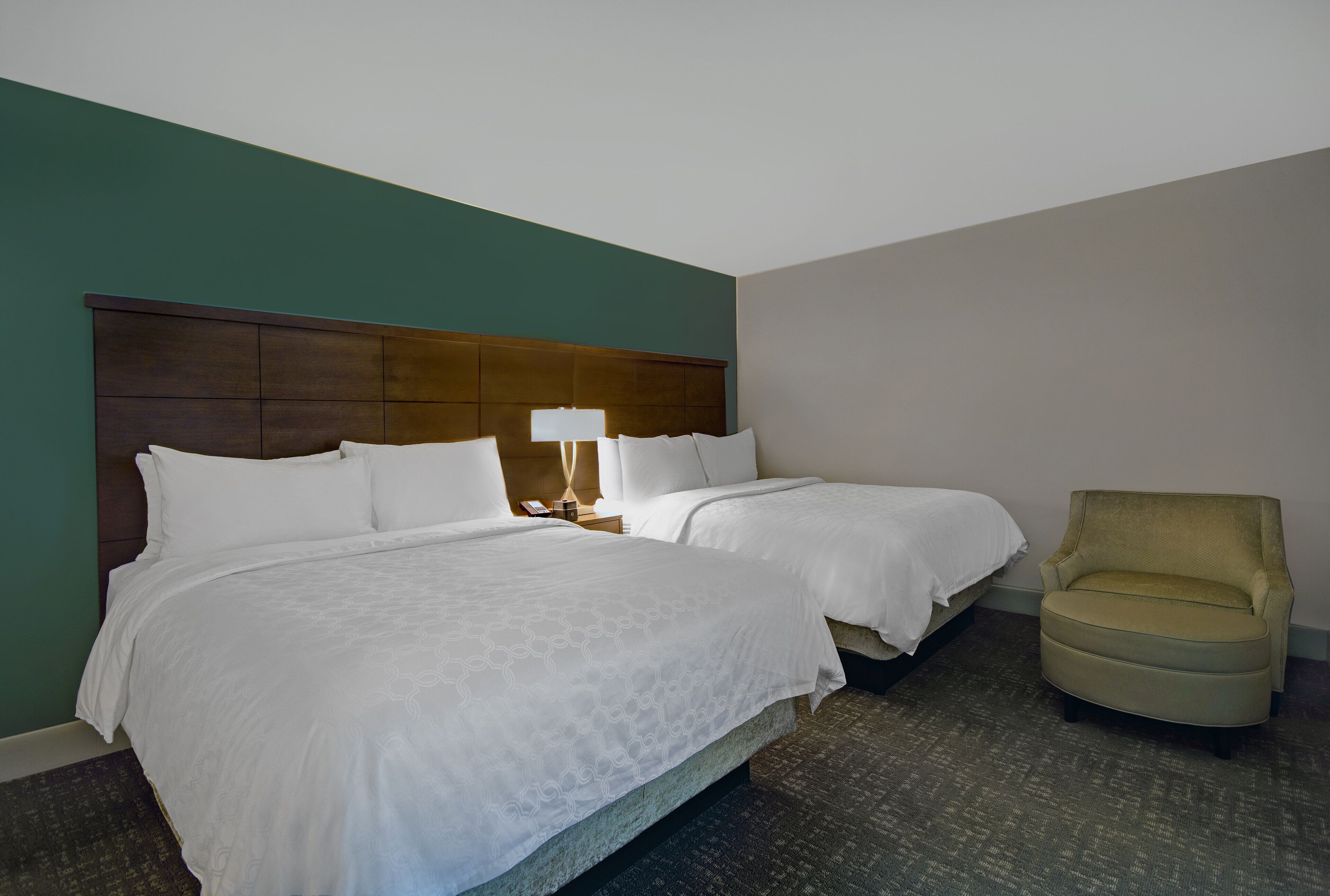 A hotel room with two beds, a nightstand with a lamp, and a chair.