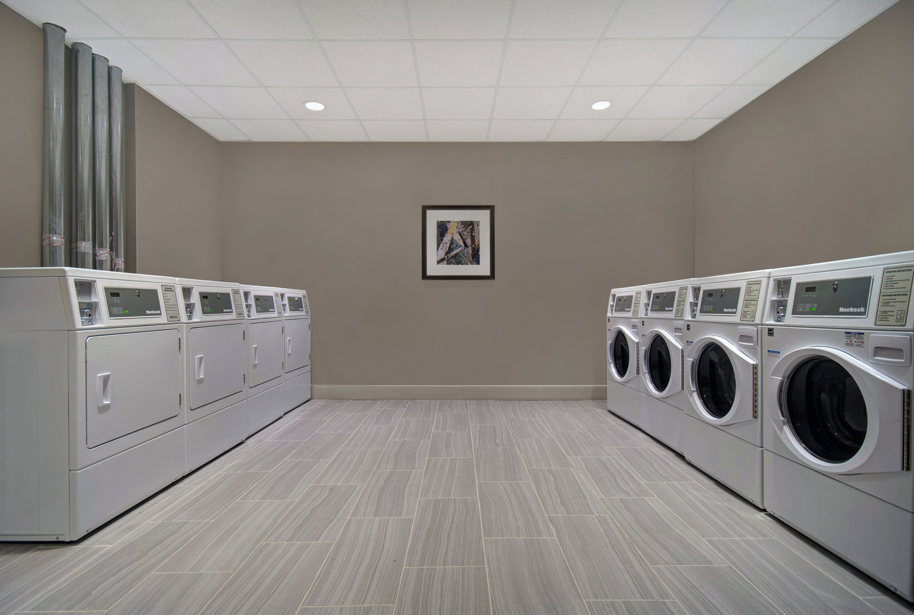 A laundry facility with rows of Bosch and Huebsch washing machines, a framed artwork on the wall, and a tiled floor.