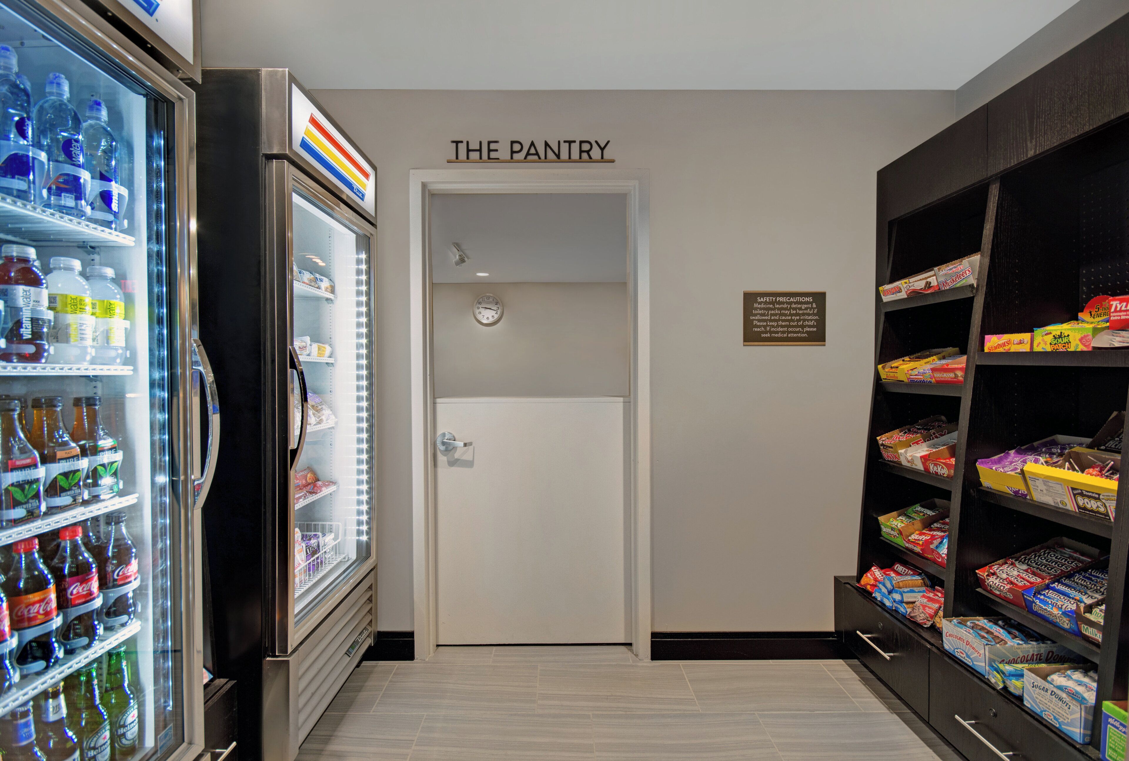 A pantry area with a refrigerator stocked with beverages, a clock on the wall, and shelves with snacks and groceries.