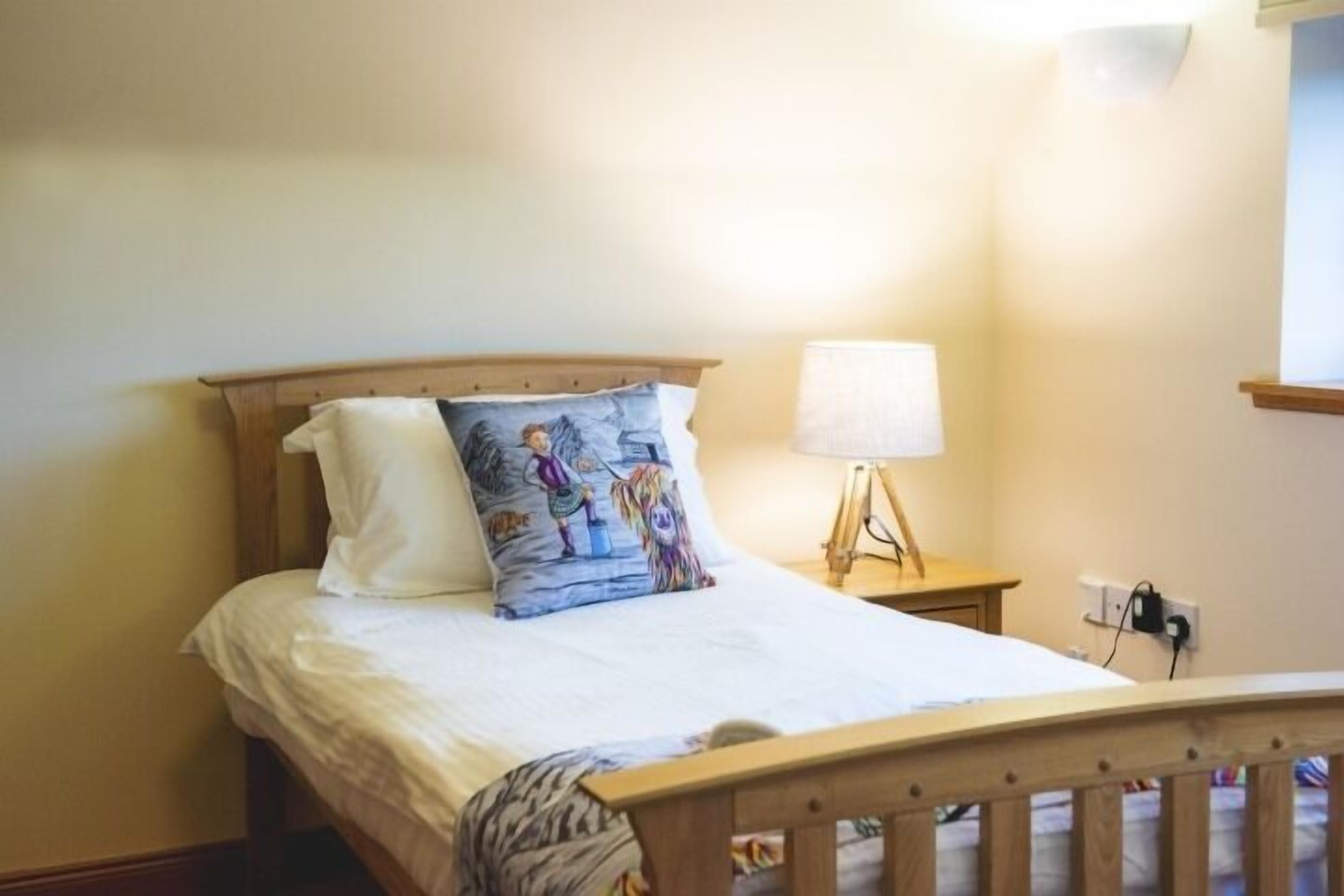 A neatly made bed with a white comforter and two pillows, one plain and one with a colorful design. There is a bedside lamp on a wooden nightstand.
