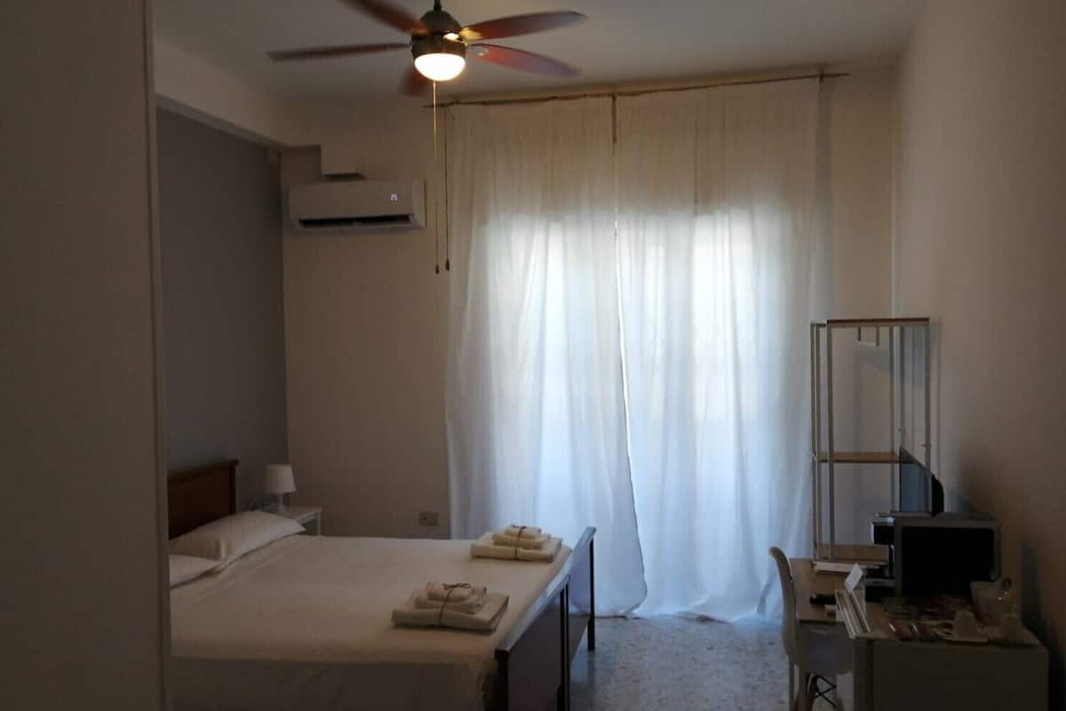 A hotel room with a bed, a desk, a chair, a ceiling fan, and a window with curtains.