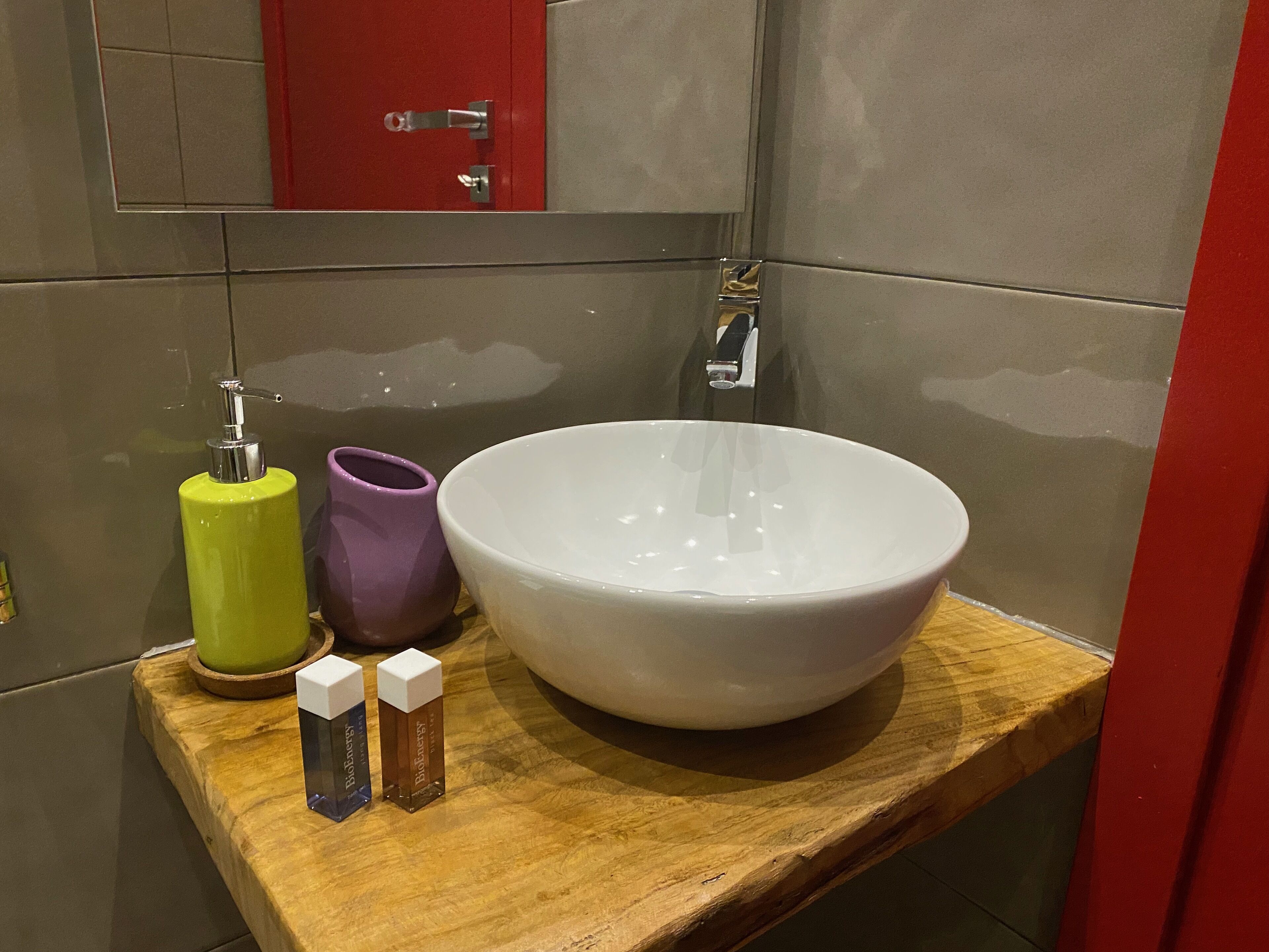 A bathroom with a wooden vanity, a white basin, a green and purple soap dispenser, and a BioEnergy soap bottle.