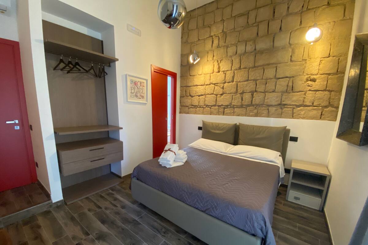 A bedroom with a bed, a wardrobe, a red door, and a stone wall.