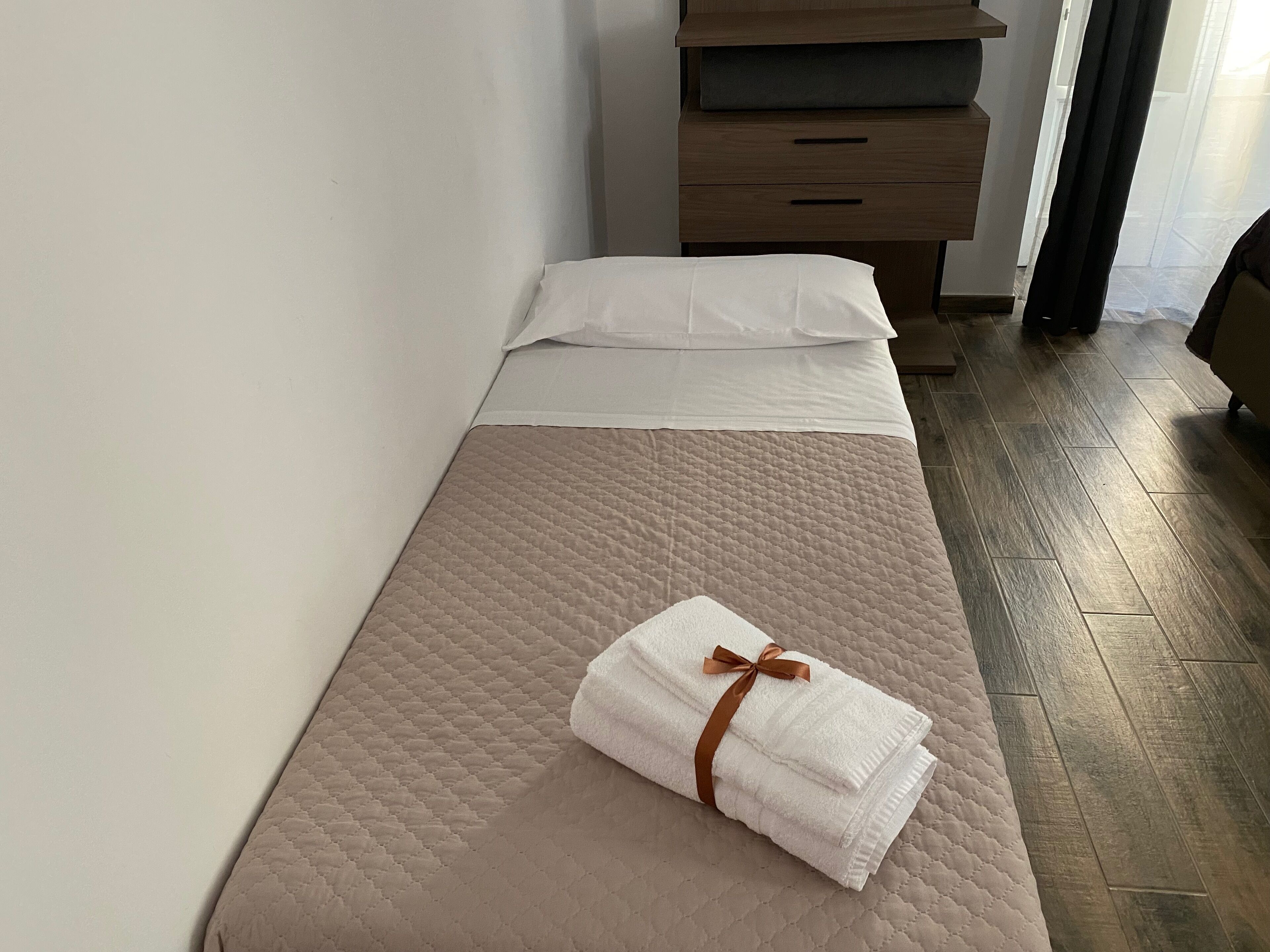 Una cama bien tendida con colcha color beige, almohadas blancas y una toalla doblada con un lazo marrón.