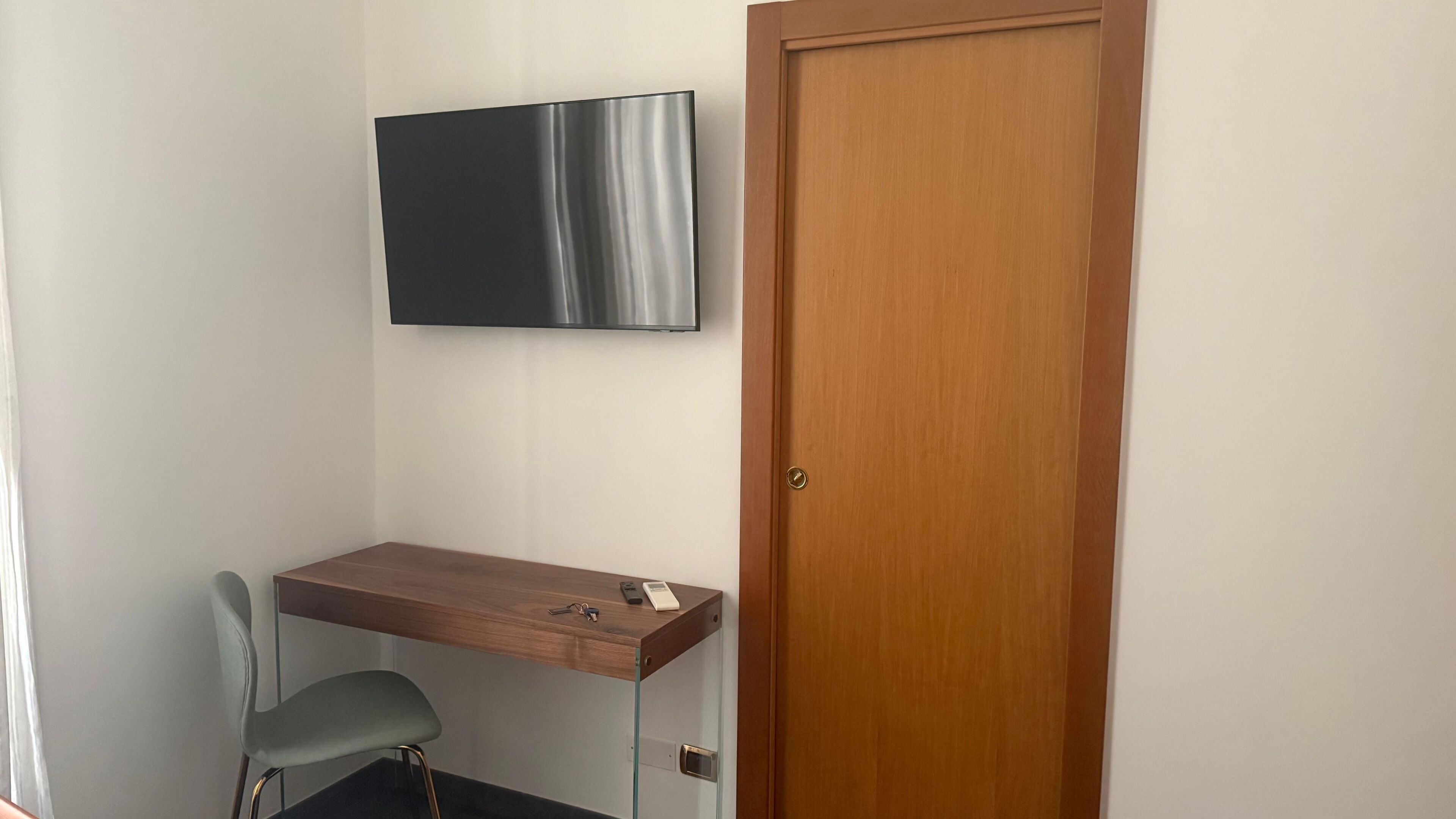 Una habitación con un escritorio de madera, una silla, un televisor montado en la pared y una puerta de madera.
