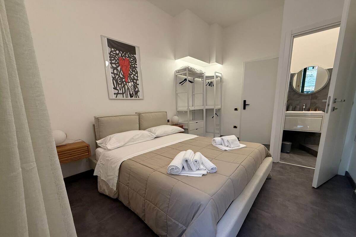 A modern hotel room with a large bed, a bedside table, a wardrobe, and a bathroom visible through an open door.