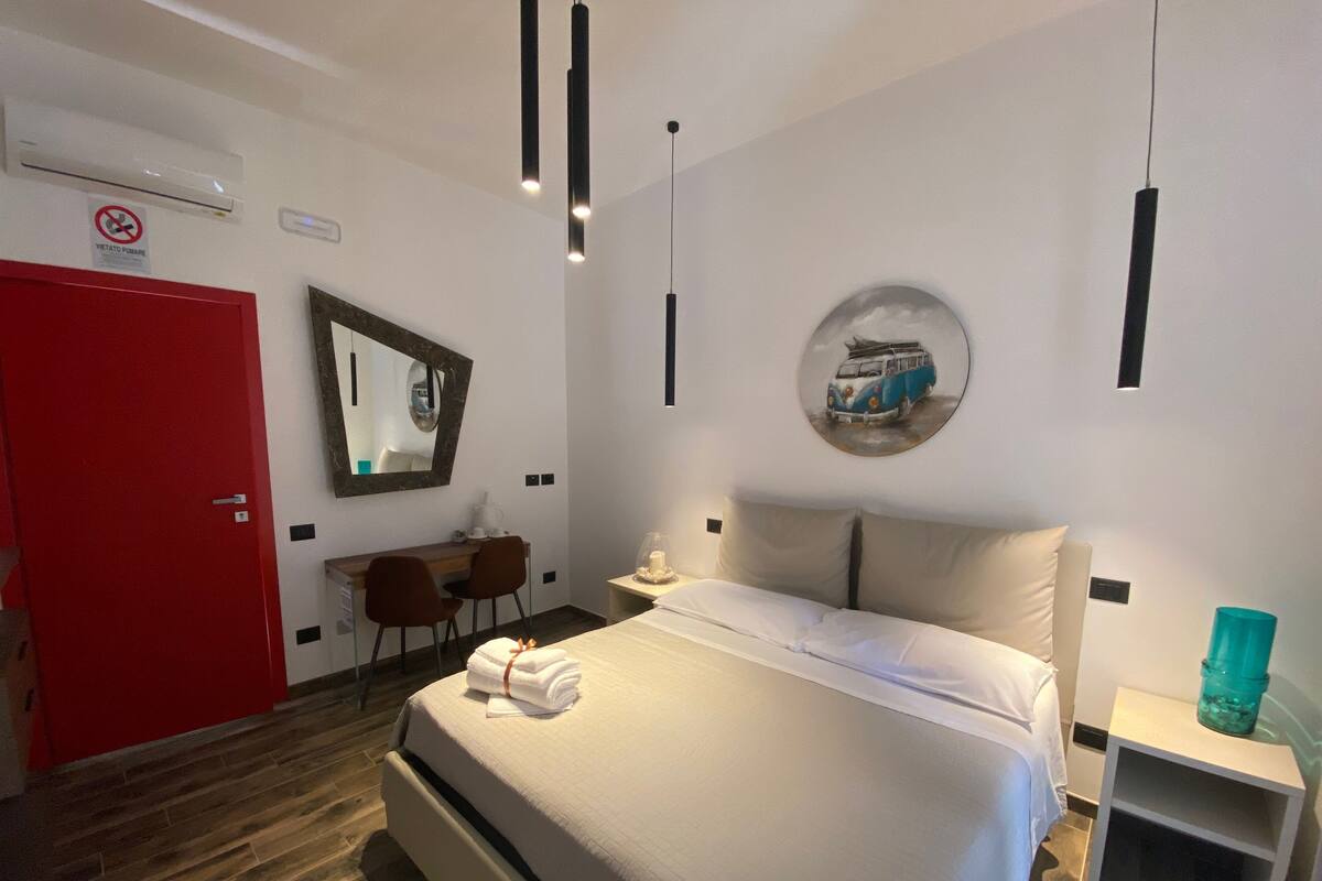 A hotel room with a bed, a desk, a chair, a red door, a mirror, and a wall-mounted picture.