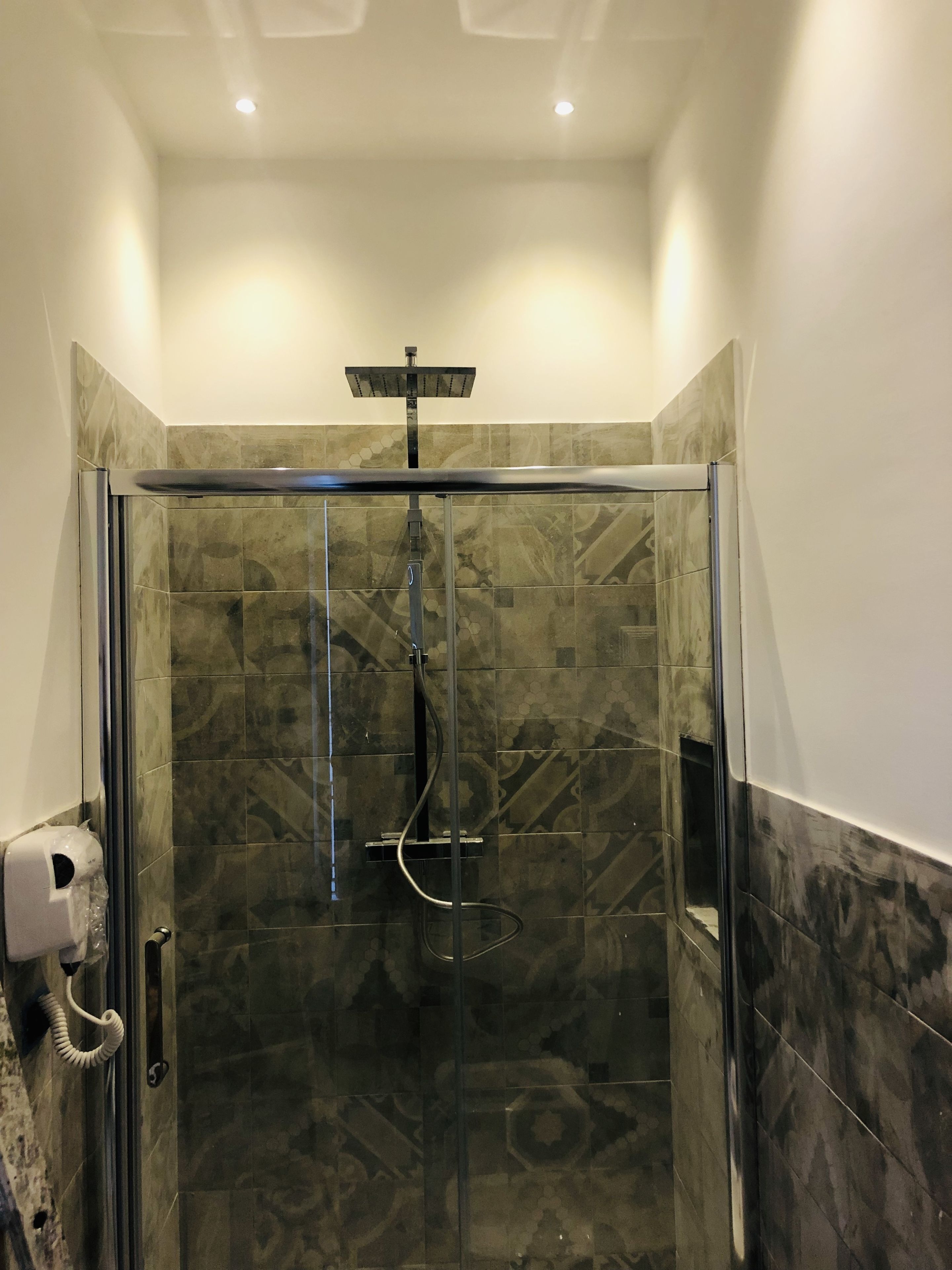 A glass-enclosed shower with a handheld showerhead, a wall-mounted control panel, and a hand dryer.