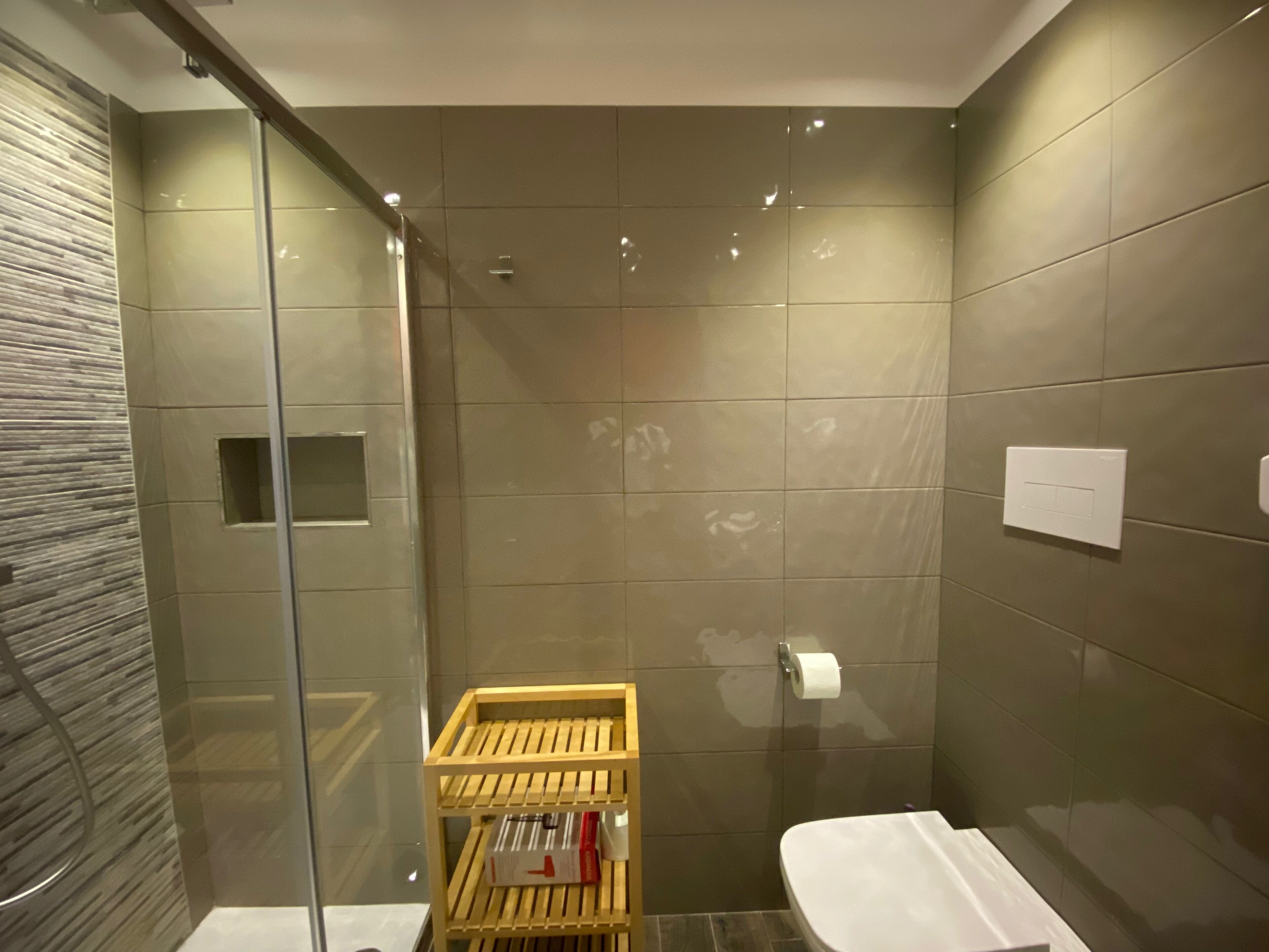 A modern bathroom with a glass shower enclosure, a wooden shelf with toiletries, and a white toilet.