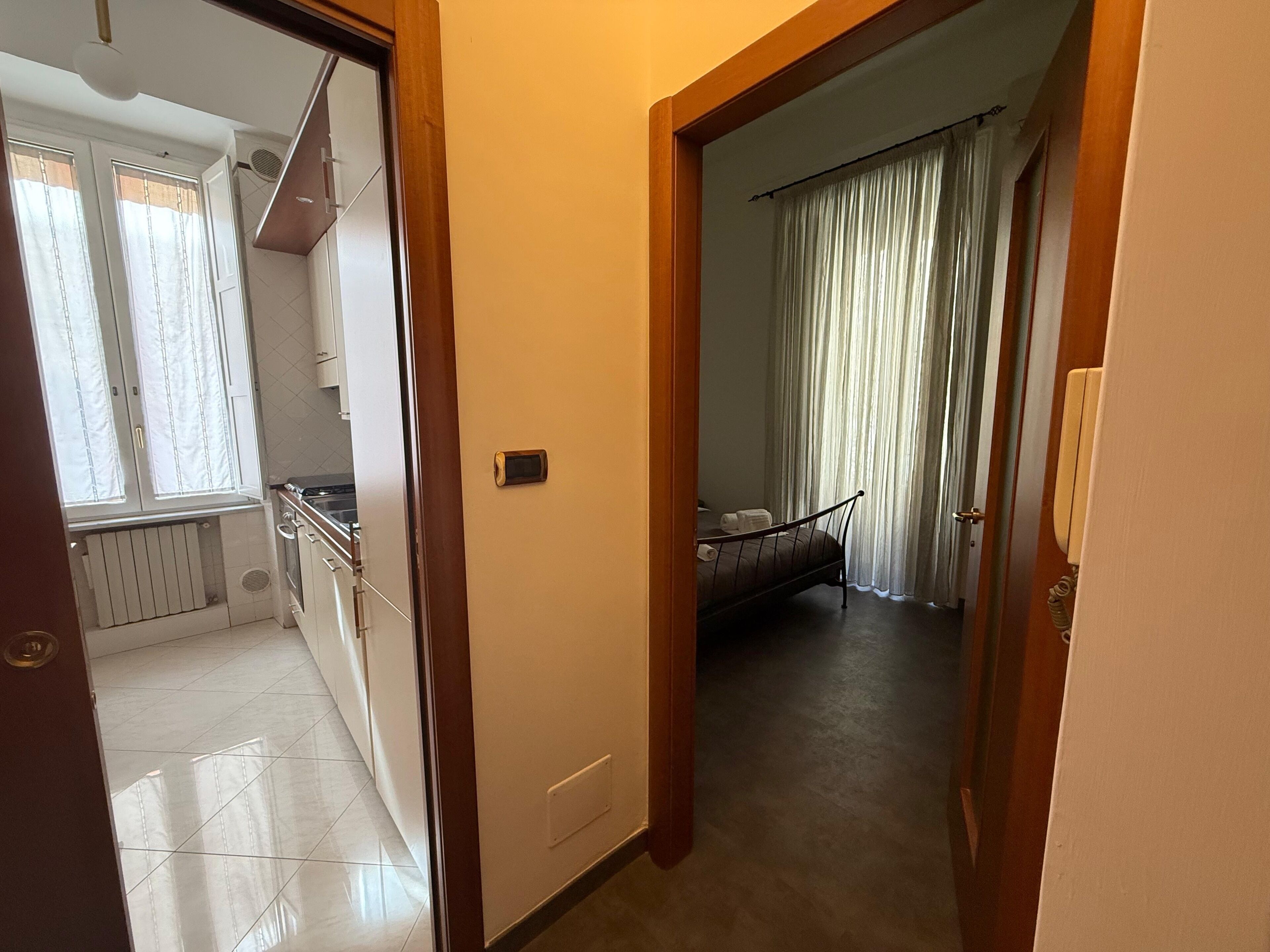 A narrow hallway leading to a bedroom with a bed, a bedside telephone, and a window with curtains.