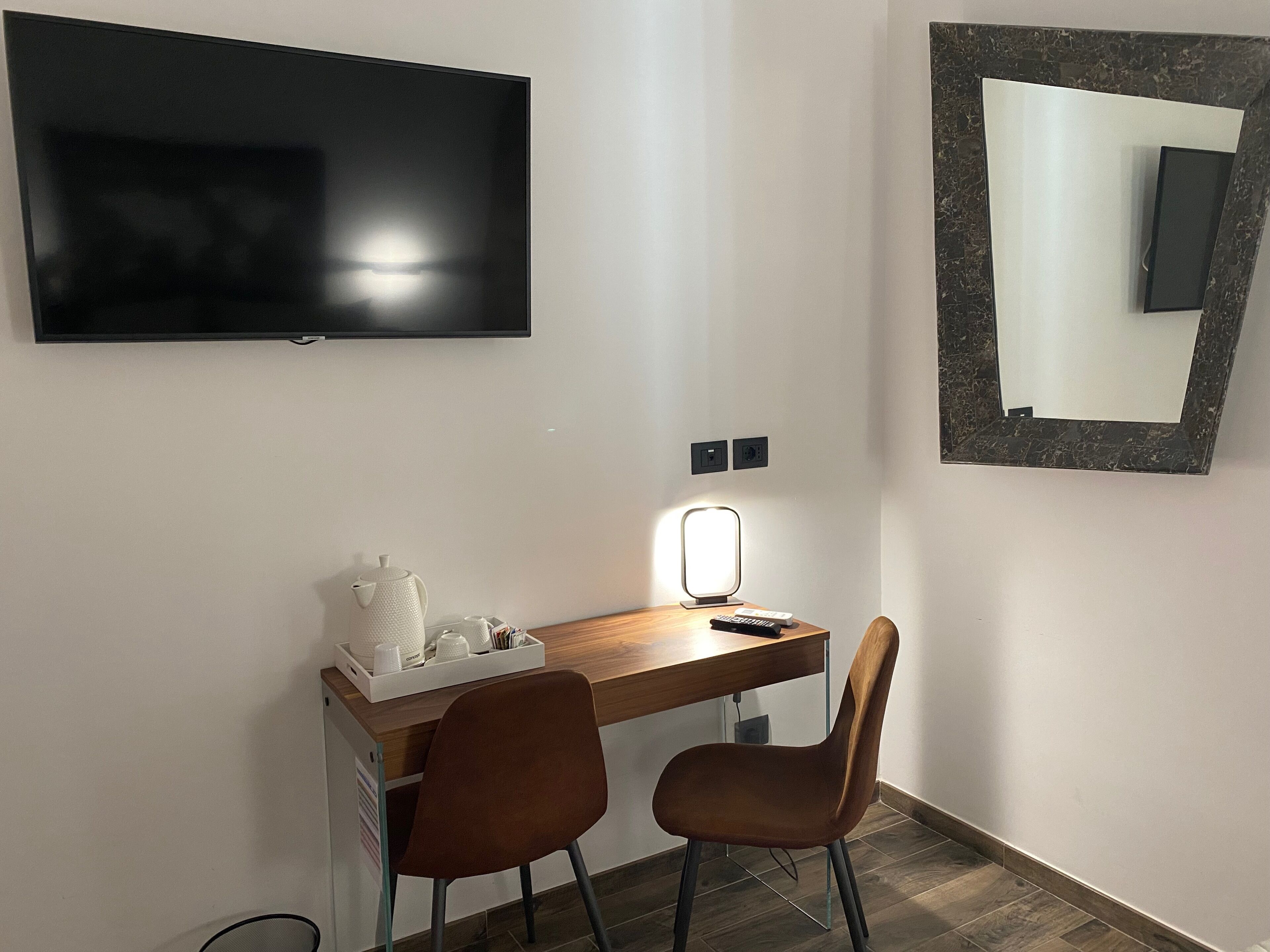 A modern room with a flat-screen TV mounted on the wall, a desk with a lamp, two chairs, and a mirror with a decorative frame.